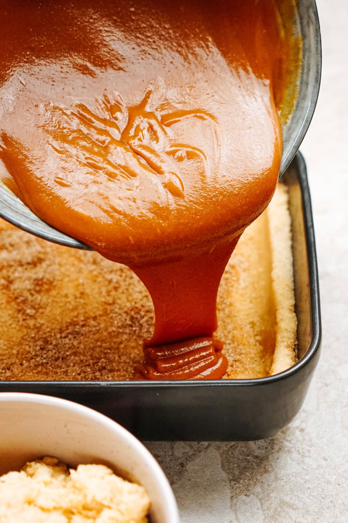 Caramel sauce being poured over a dessert crust in a baking pan, with a bowl of ingredients in the foreground.