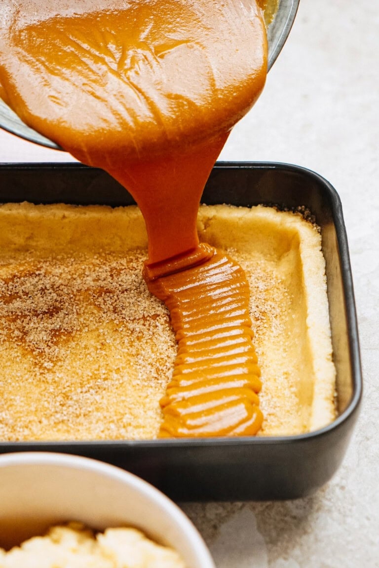 Caramel sauce is being poured into a baking pan lined with dough and sprinkled with sugar.