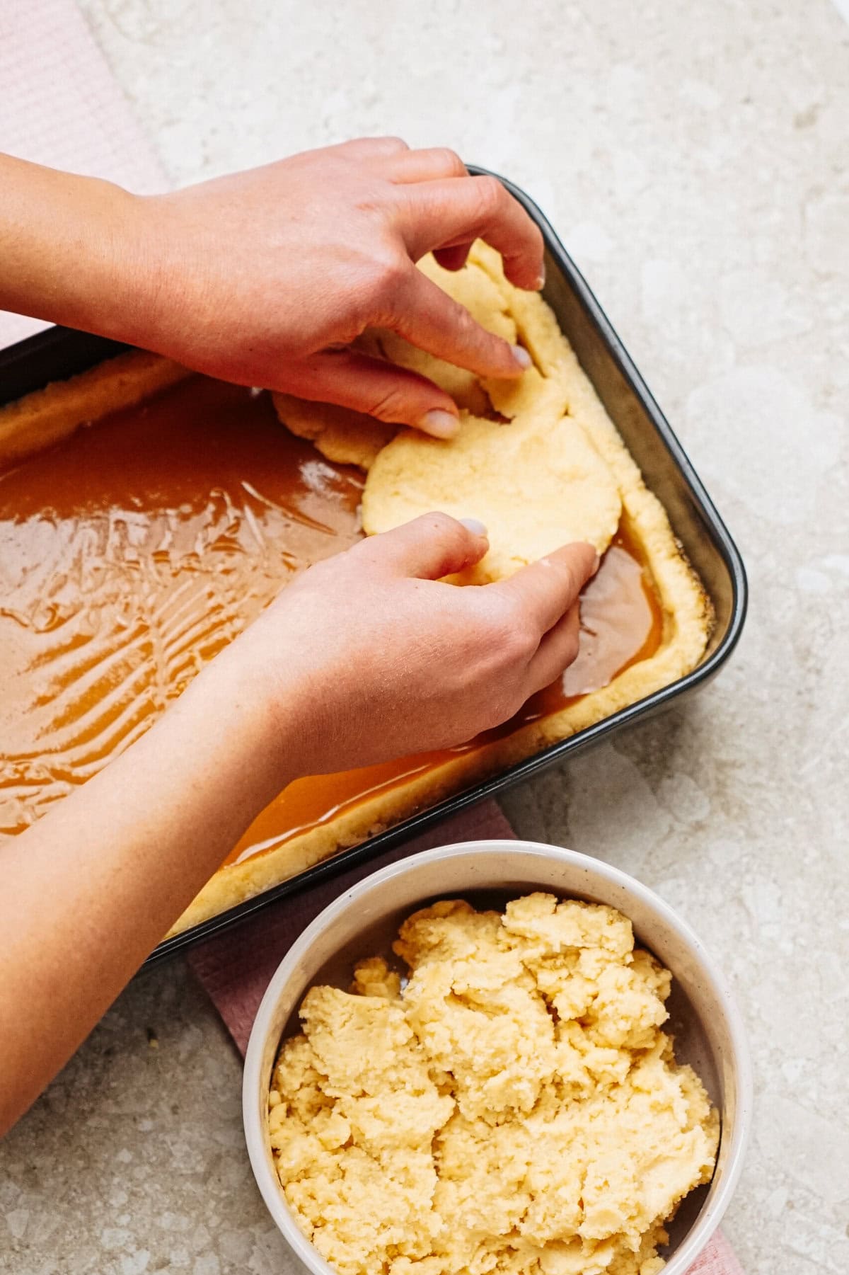 Hands pressing dough onto a sheet of caramel in a baking pan, with a bowl of extra dough nearby on a countertop.