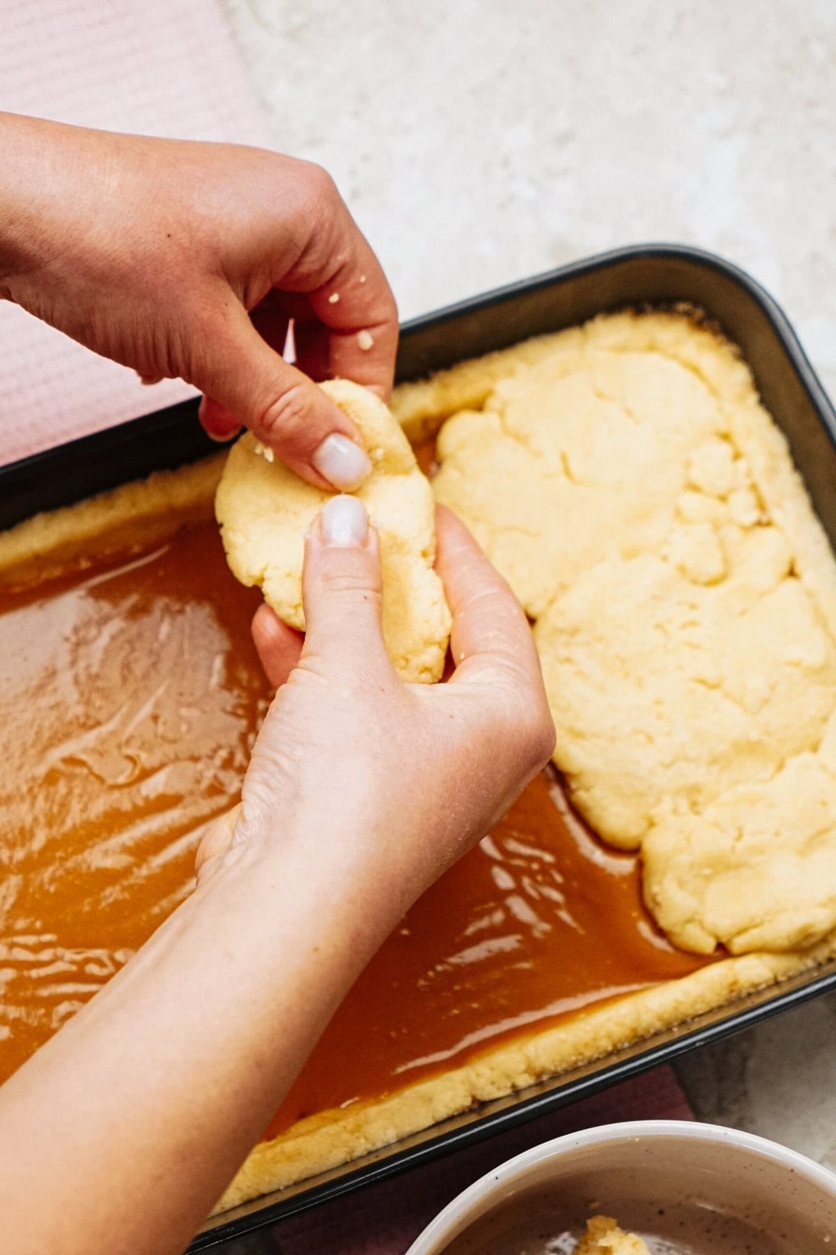 Hands pressing dough onto a caramel-filled baking dish.