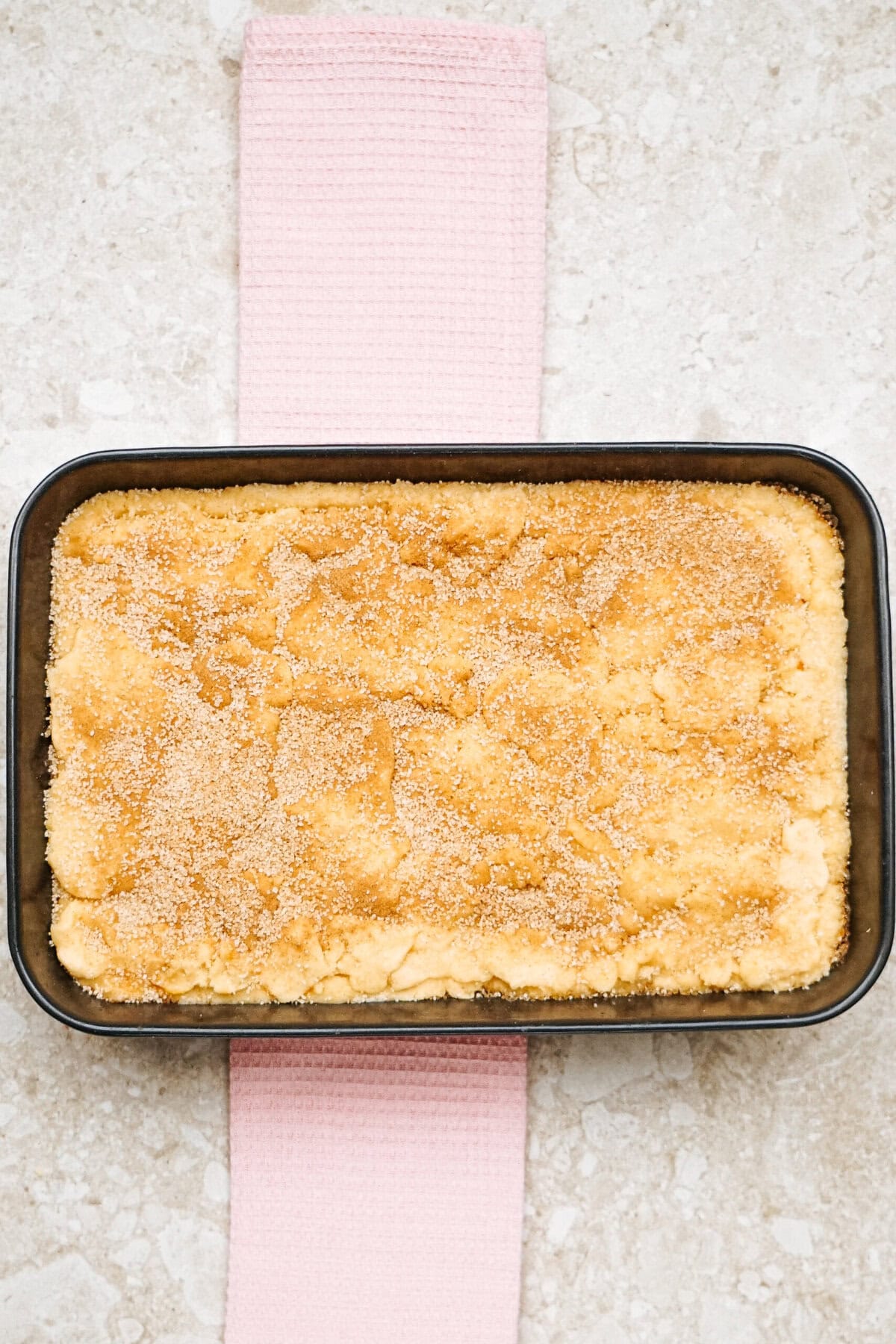 A rectangular baking dish filled with crumb-topped baked dessert rests on a pink cloth on a light stone surface.