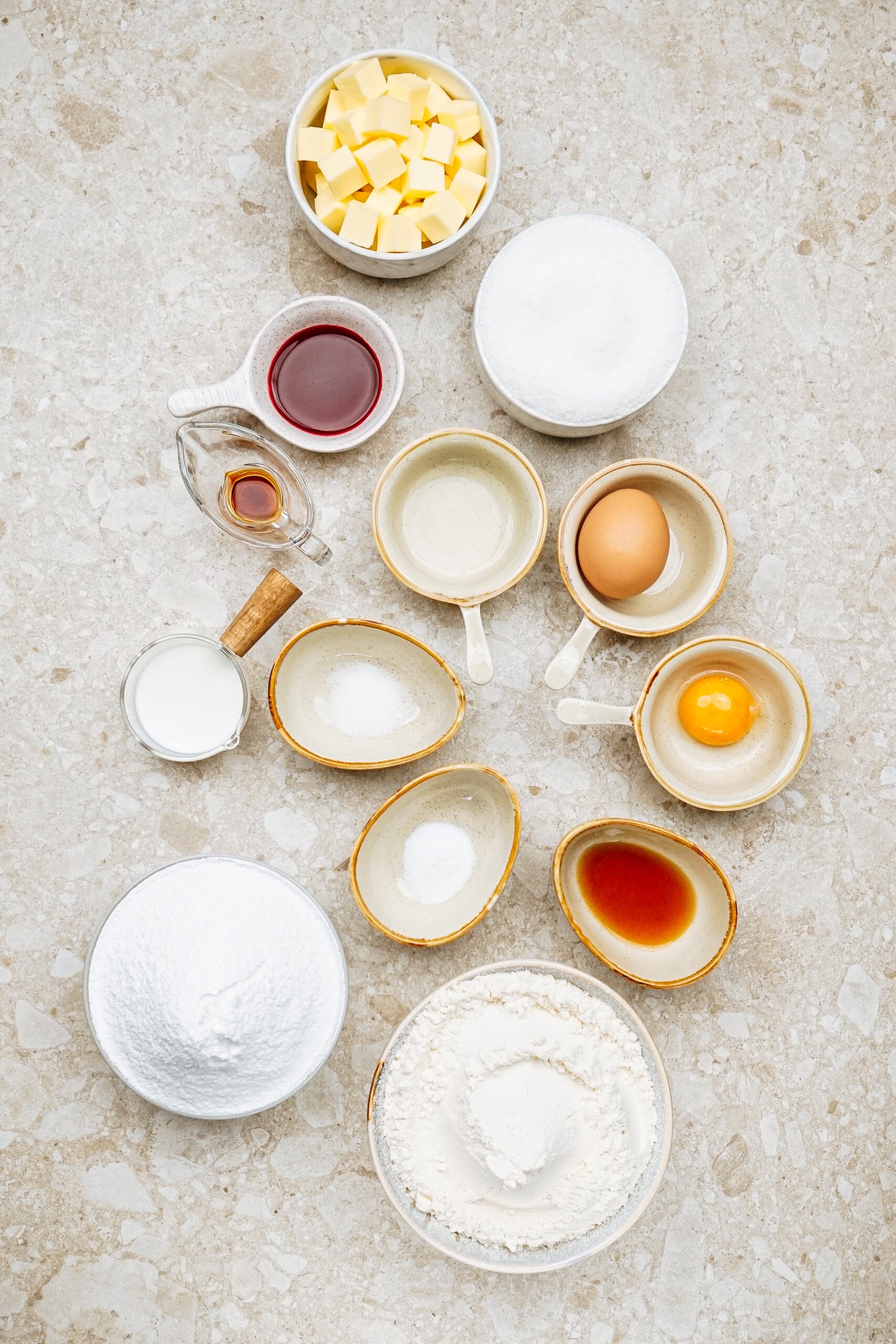 Assorted baking ingredients: butter cubes, sugar, eggs, vanilla extract, milk, flour, baking soda, baking powder, and salt in bowls on a textured surface.