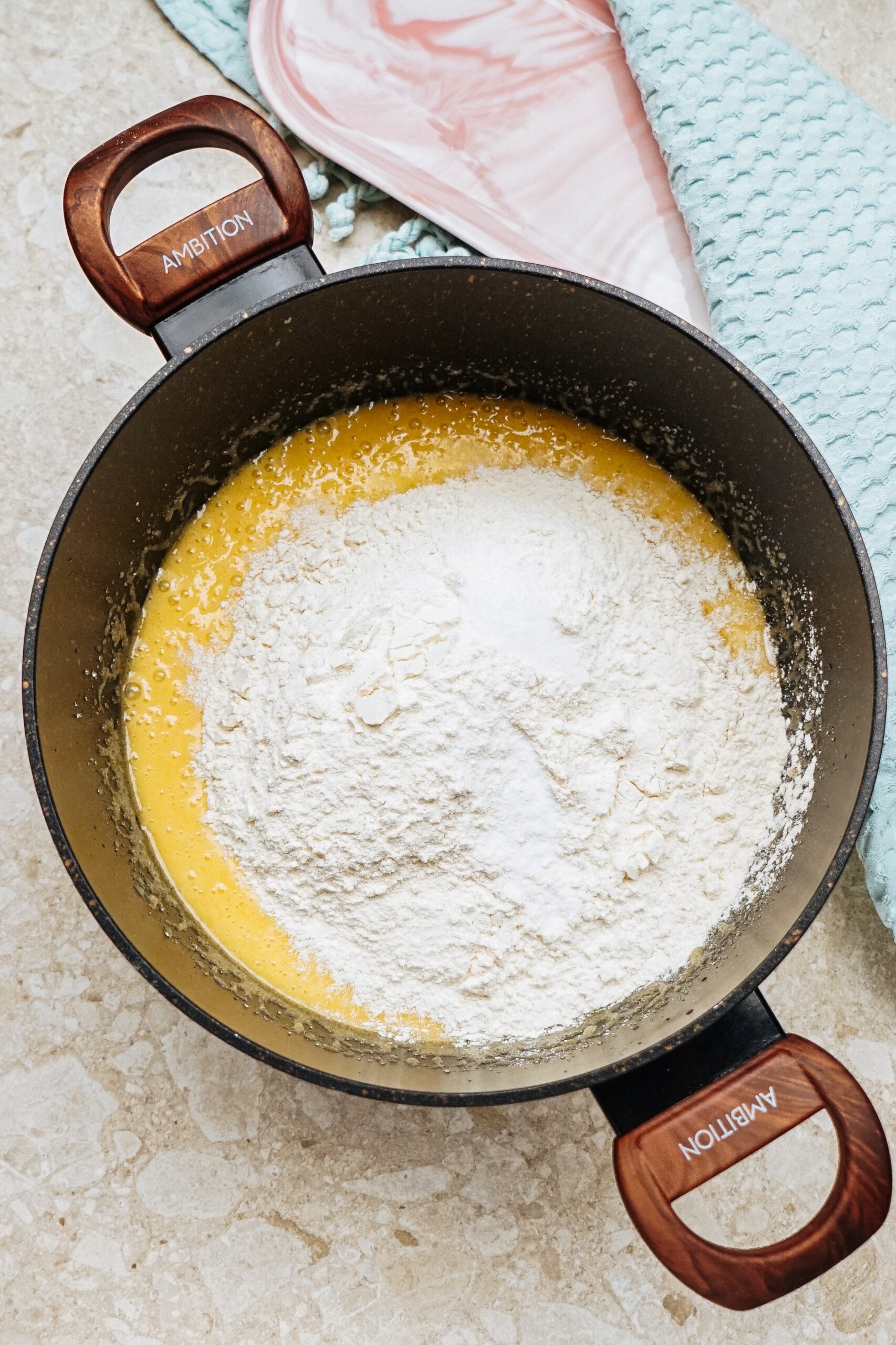 A mixing bowl with cookie batter, featuring flour on top. Two wooden-handled spatulas are visible nearby on a textured countertop.