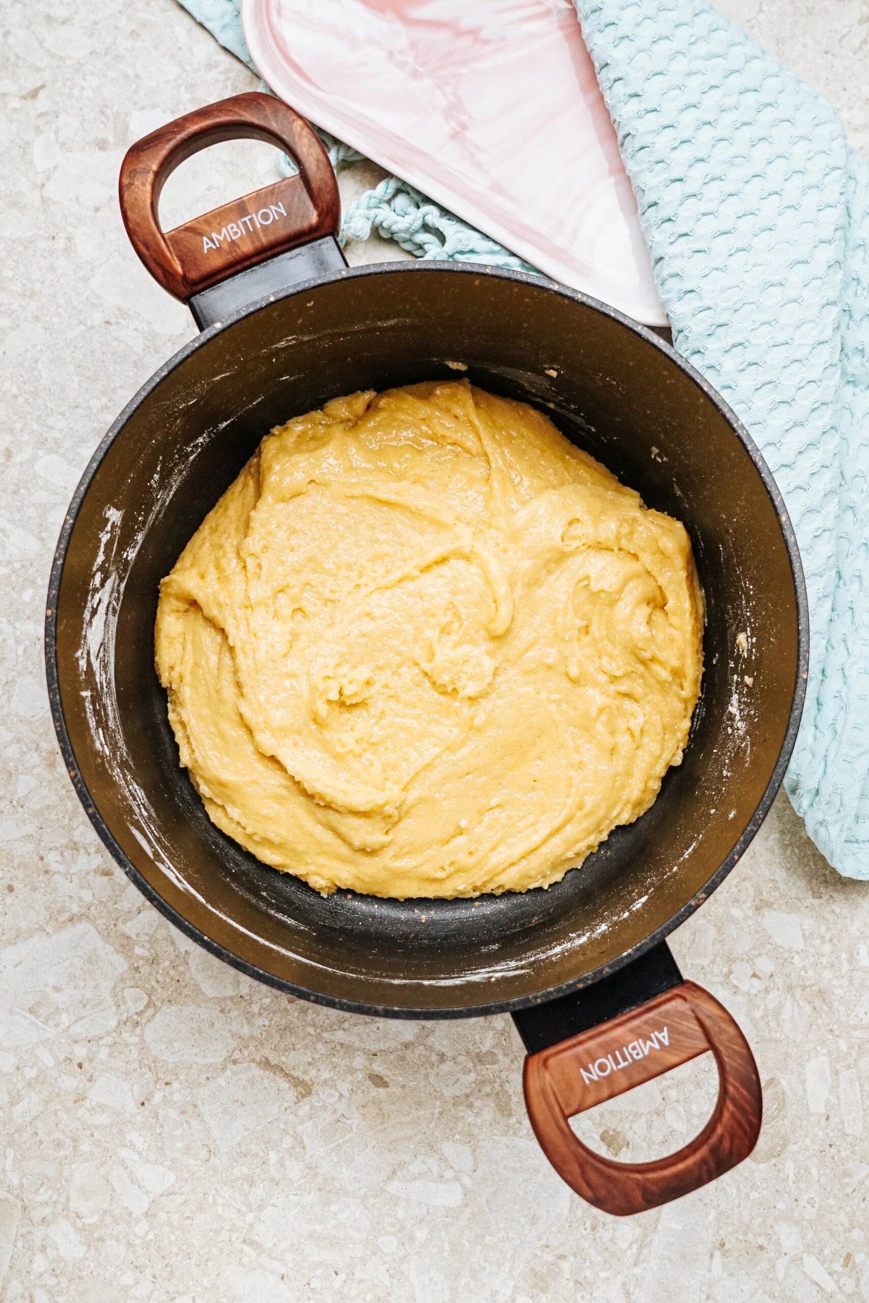 A pot containing a creamy, yellow batter sits on a light stone countertop, next to a blue textured cloth.