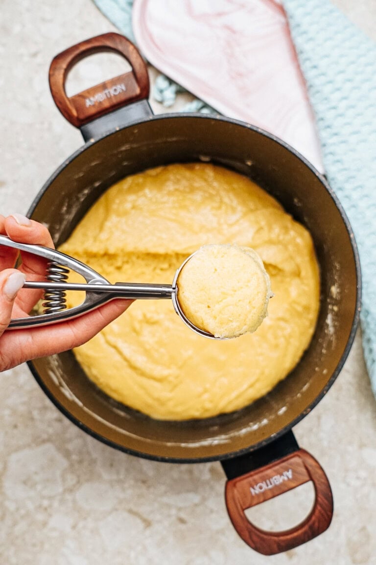 A hand holding a scoop of yellow batter above a pot filled with the same batter. Brown-handled pot on a countertop with a blue towel and a pink dish in the background.