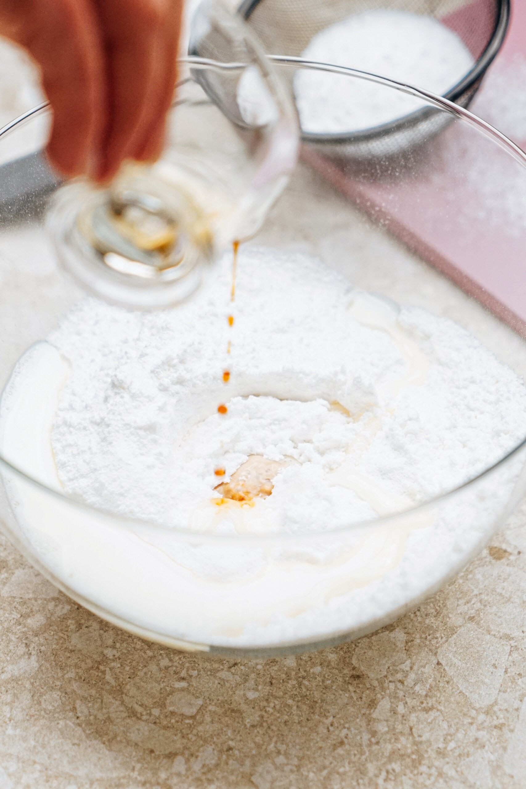 A hand pours vanilla extract into a glass bowl containing flour and other dry ingredients.