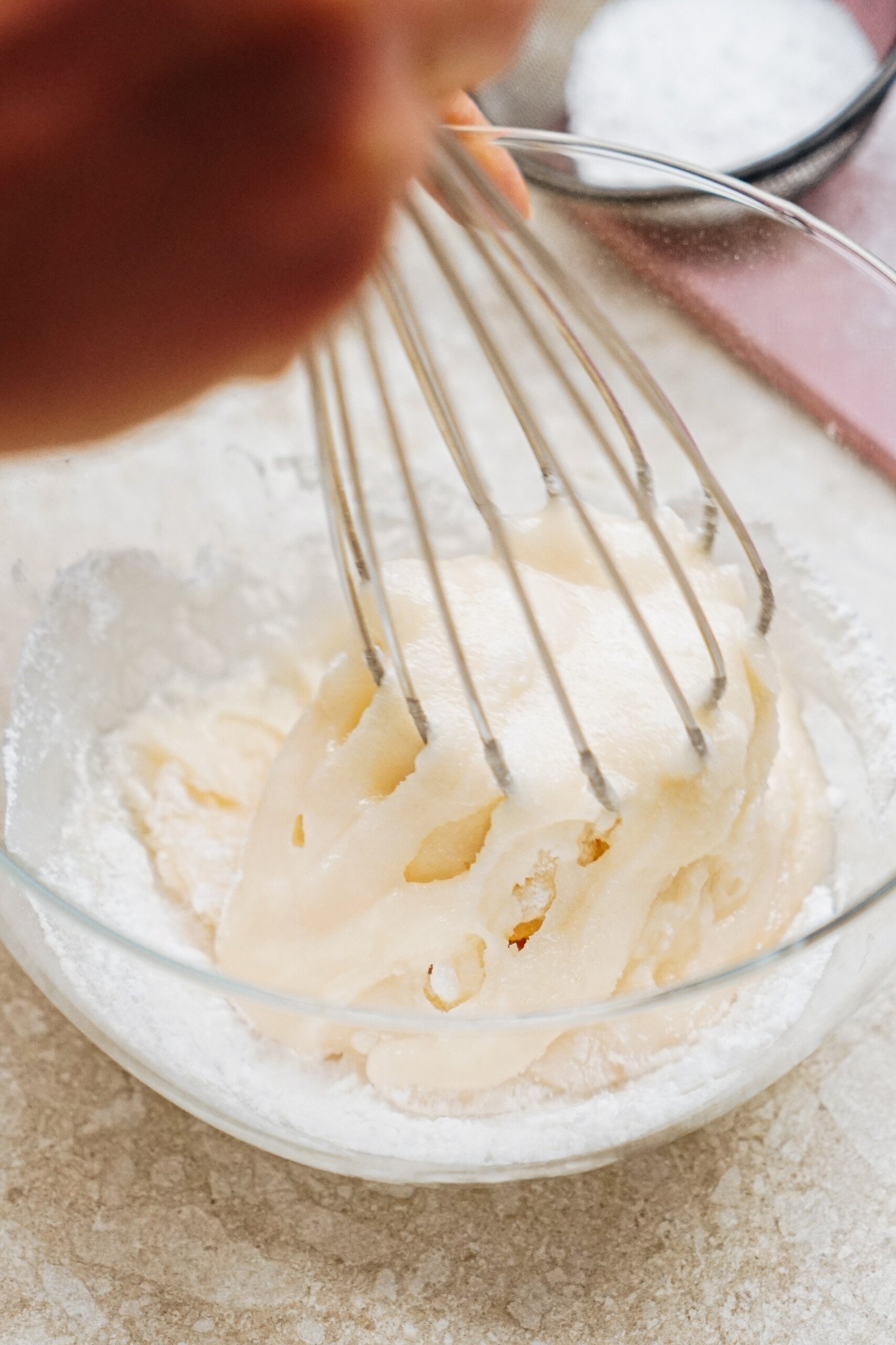 A person's hand uses a whisk to mix a creamy batter in a glass bowl on a countertop.