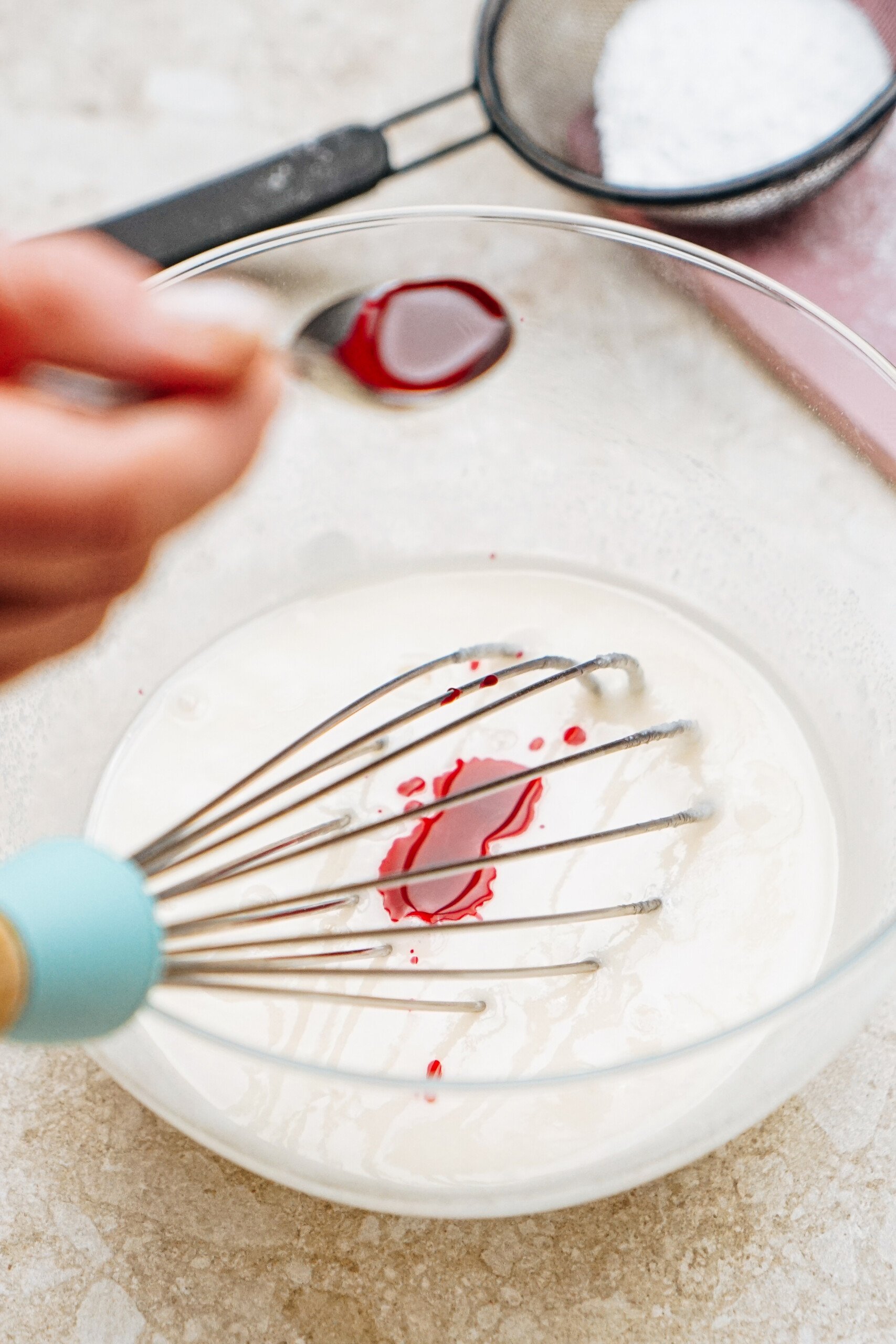 Hand holding a teaspoon adds red liquid to a glass bowl with white mixture and whisk; sifter with white powder in background.