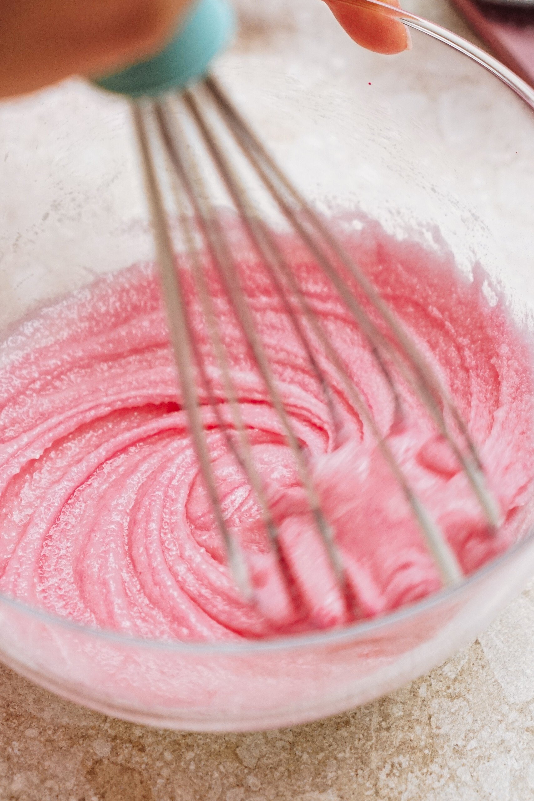 A hand whisking pink batter in a clear glass bowl on a light countertop.