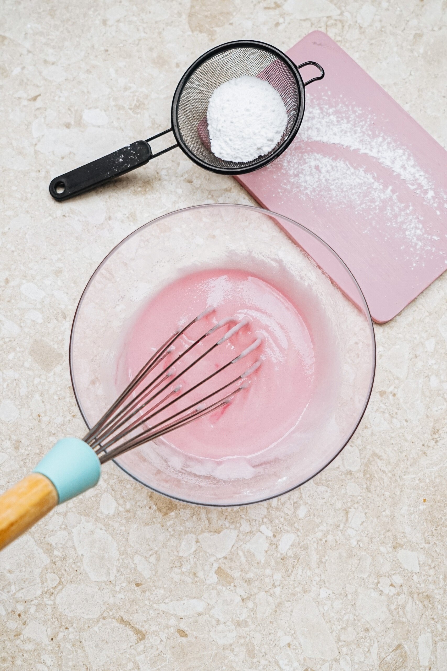 A whisk in a bowl with pink batter. A sifter with powdered sugar is nearby on a pink cutting board.