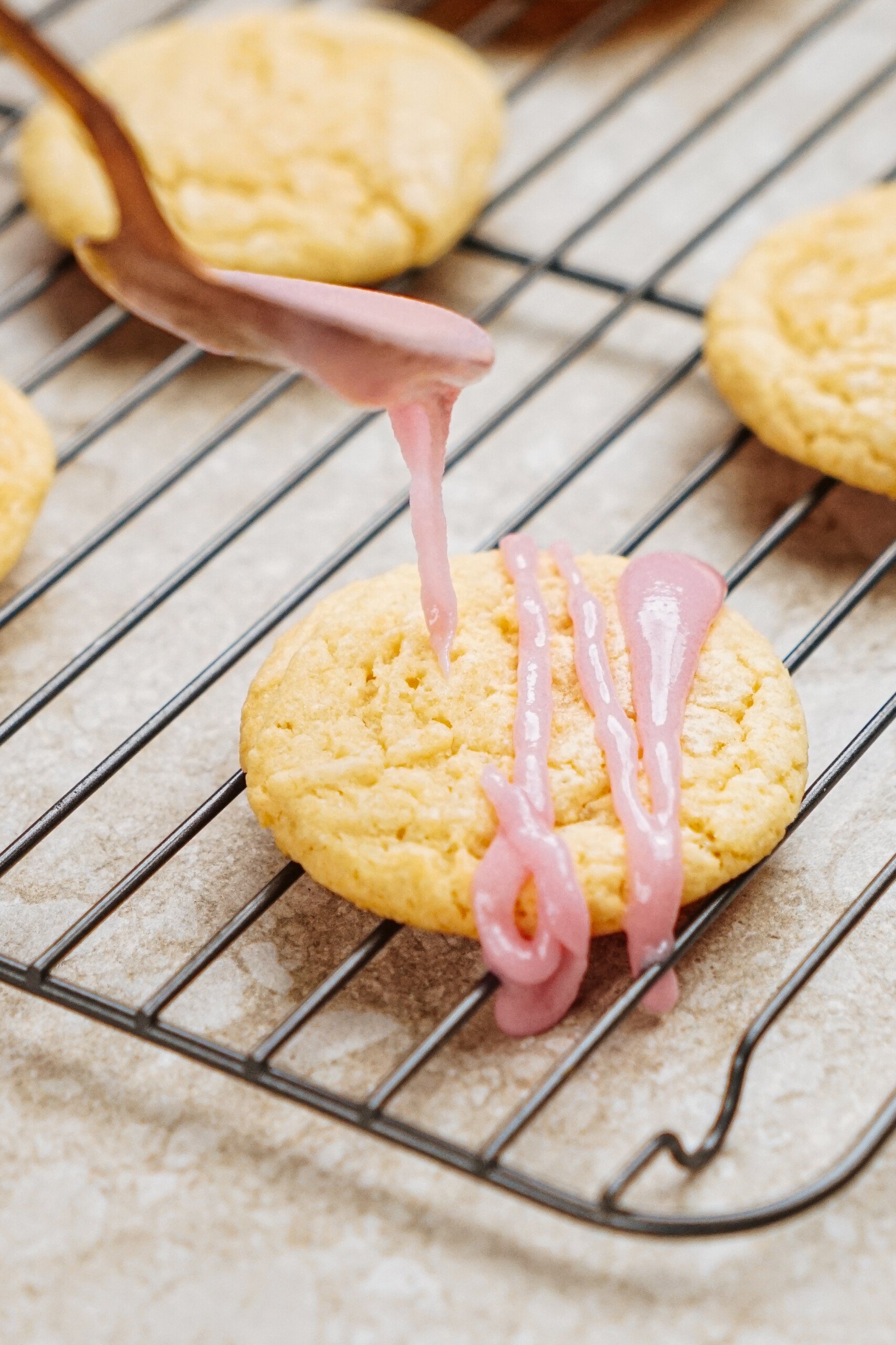 A cookie on a cooling rack being drizzled with pink icing from a spoon.