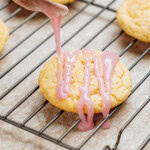 A spoon drizzles pink icing onto a round cookie resting on a cooling rack. Other cookies are visible in the background.