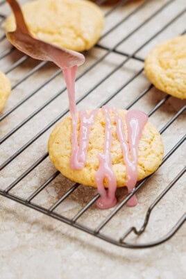 A spoon drizzles pink icing onto a round cookie resting on a cooling rack. Other cookies are visible in the background.