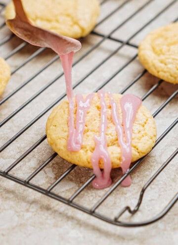 A spoon drizzles pink icing onto a round cookie resting on a cooling rack. Other cookies are visible in the background.