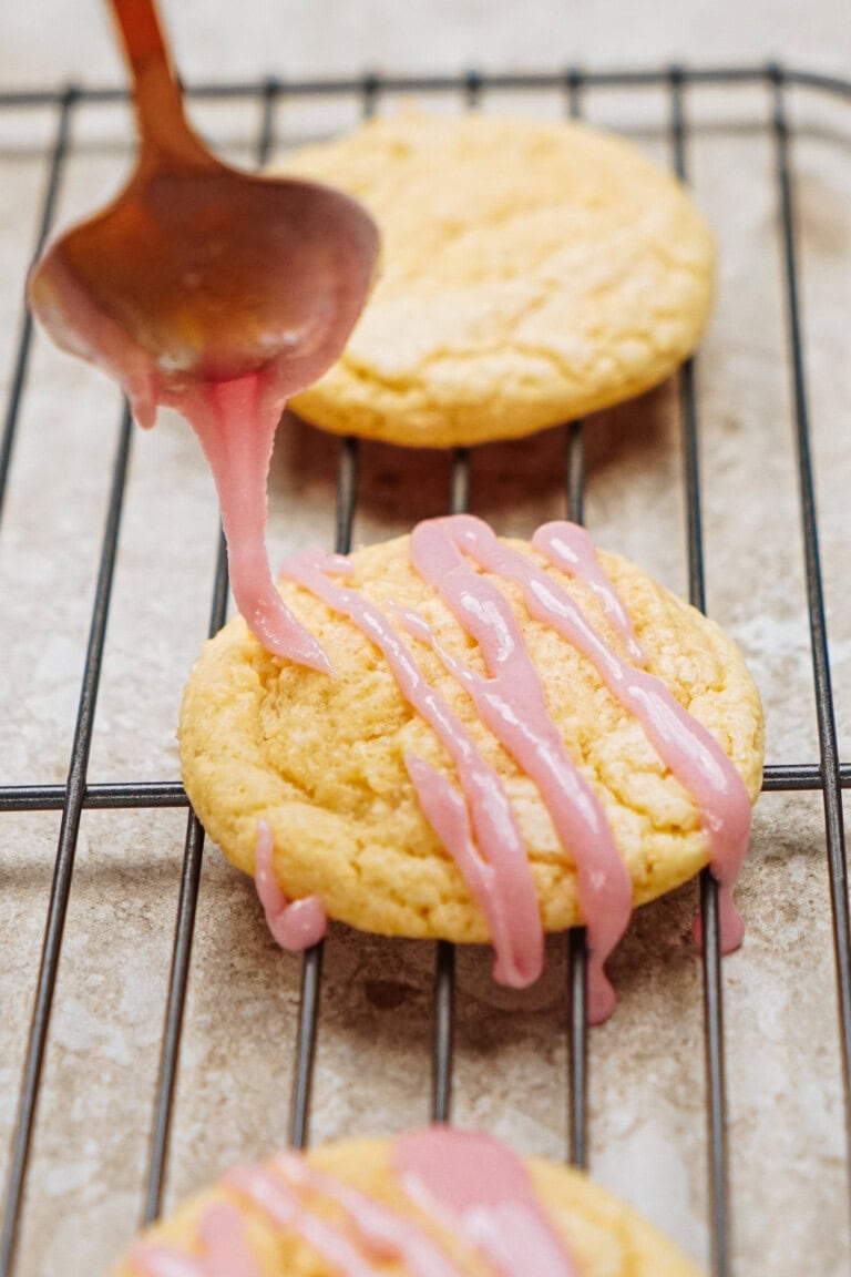 Pink icing is being drizzled onto round cookies on a cooling rack.