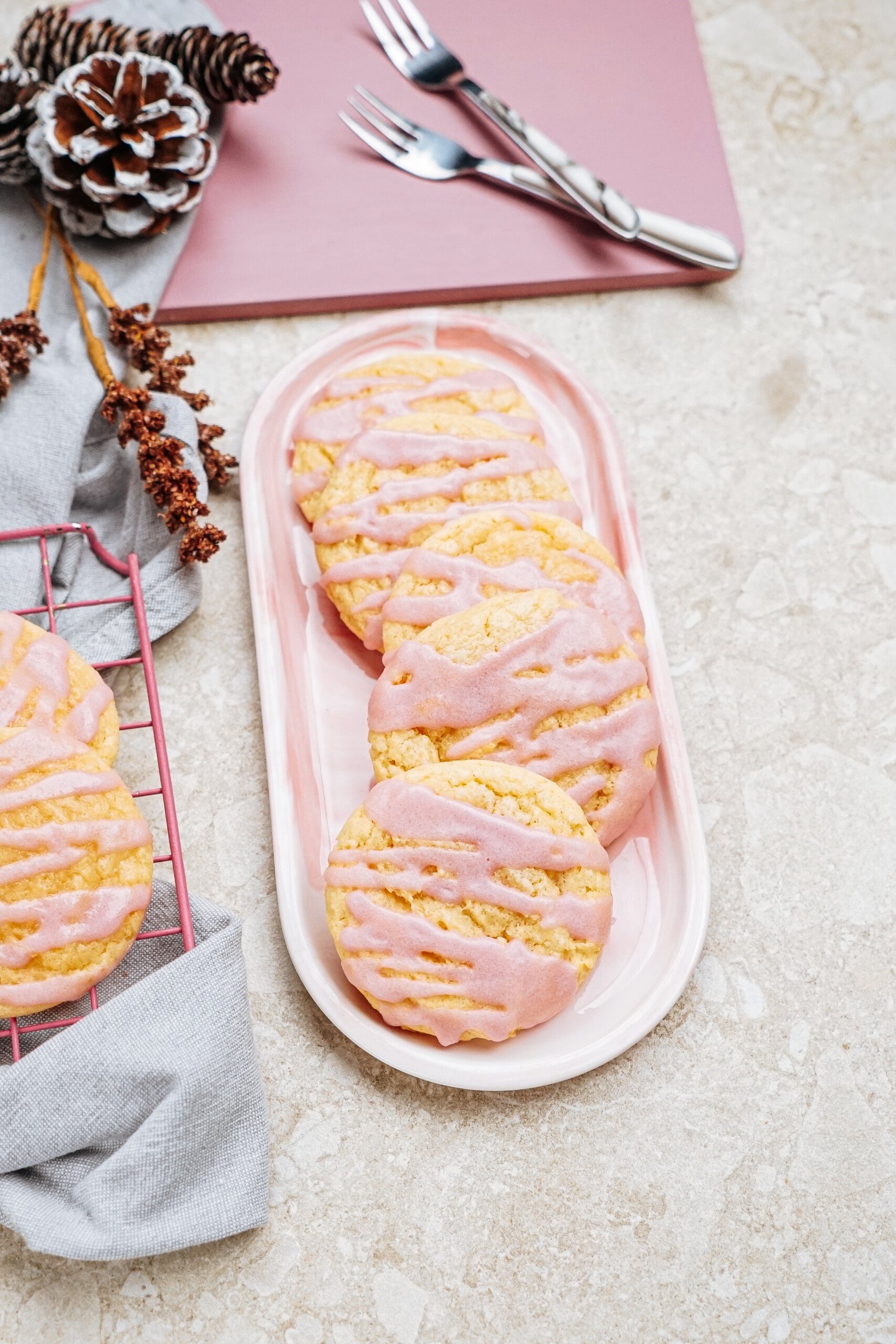 Pink glazed cookies on a rectangular plate with a pinecone and cutlery nearby, on a textured surface.