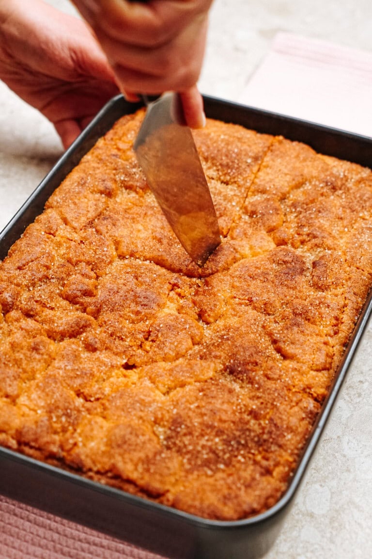 A person slices a golden-brown cake in a rectangular baking pan on a countertop.