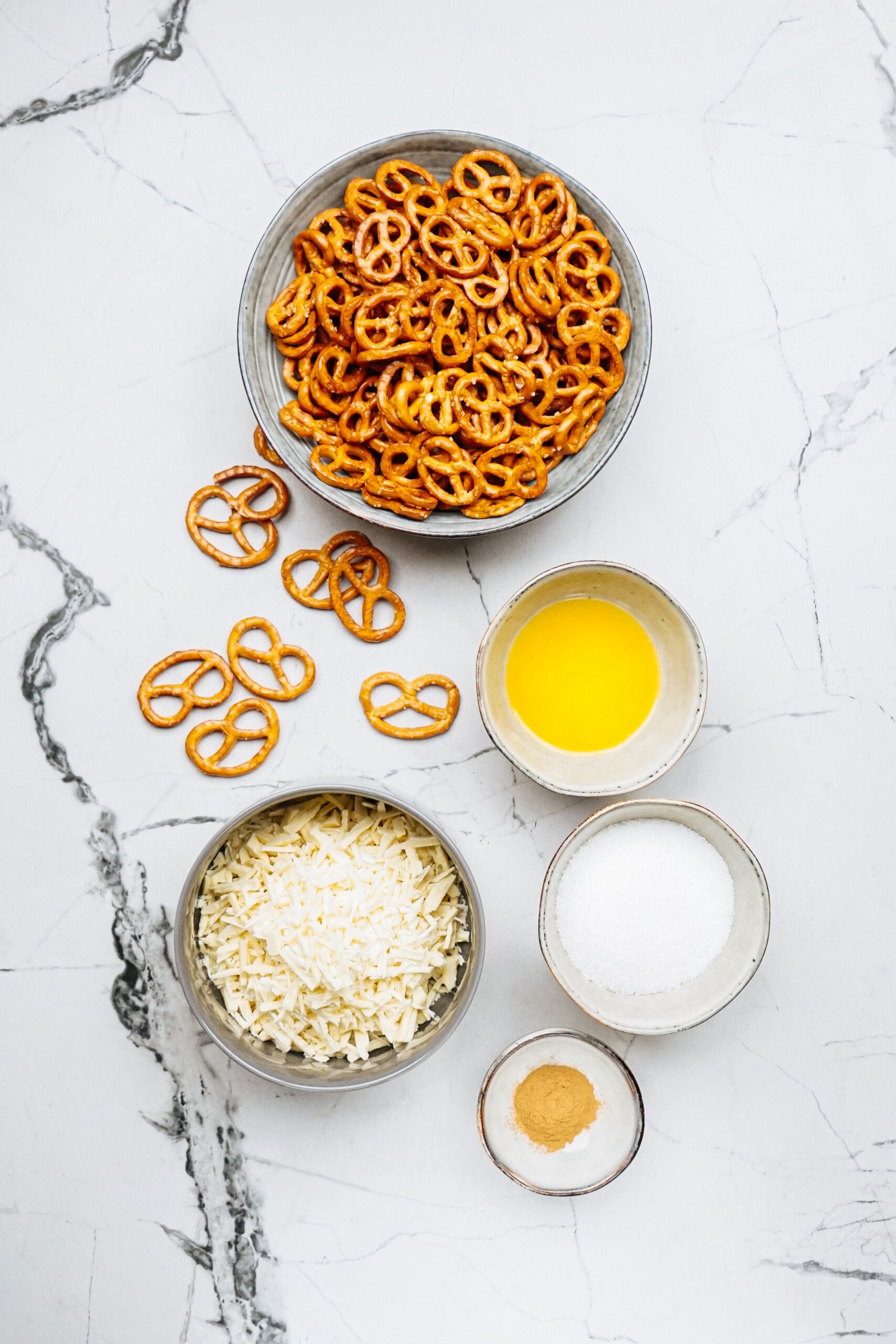 A bowl of pretzels sits invitingly alongside small bowls of melted butter, shredded cheese, sugar, and cinnamon on a marbled surface, creating the perfect setup for crafting your own cinnamon roll pretzels.