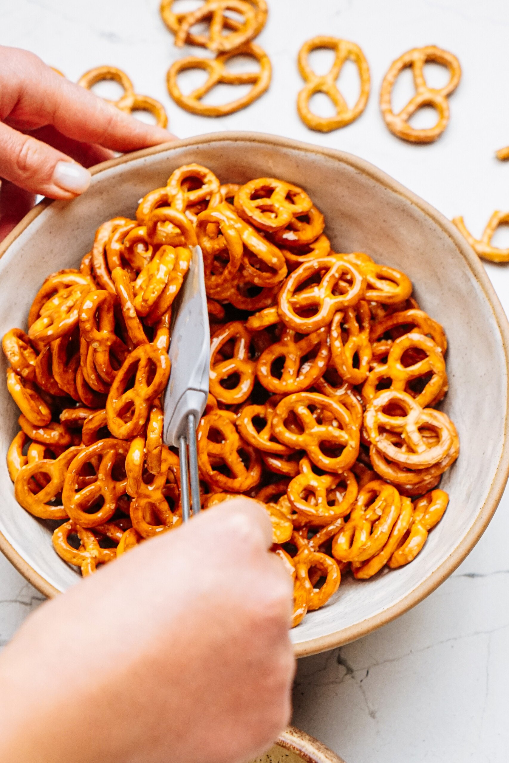 A person stirs cinnamon roll pretzels in a bowl using a knife, with additional pretzels scattered around on a white surface.