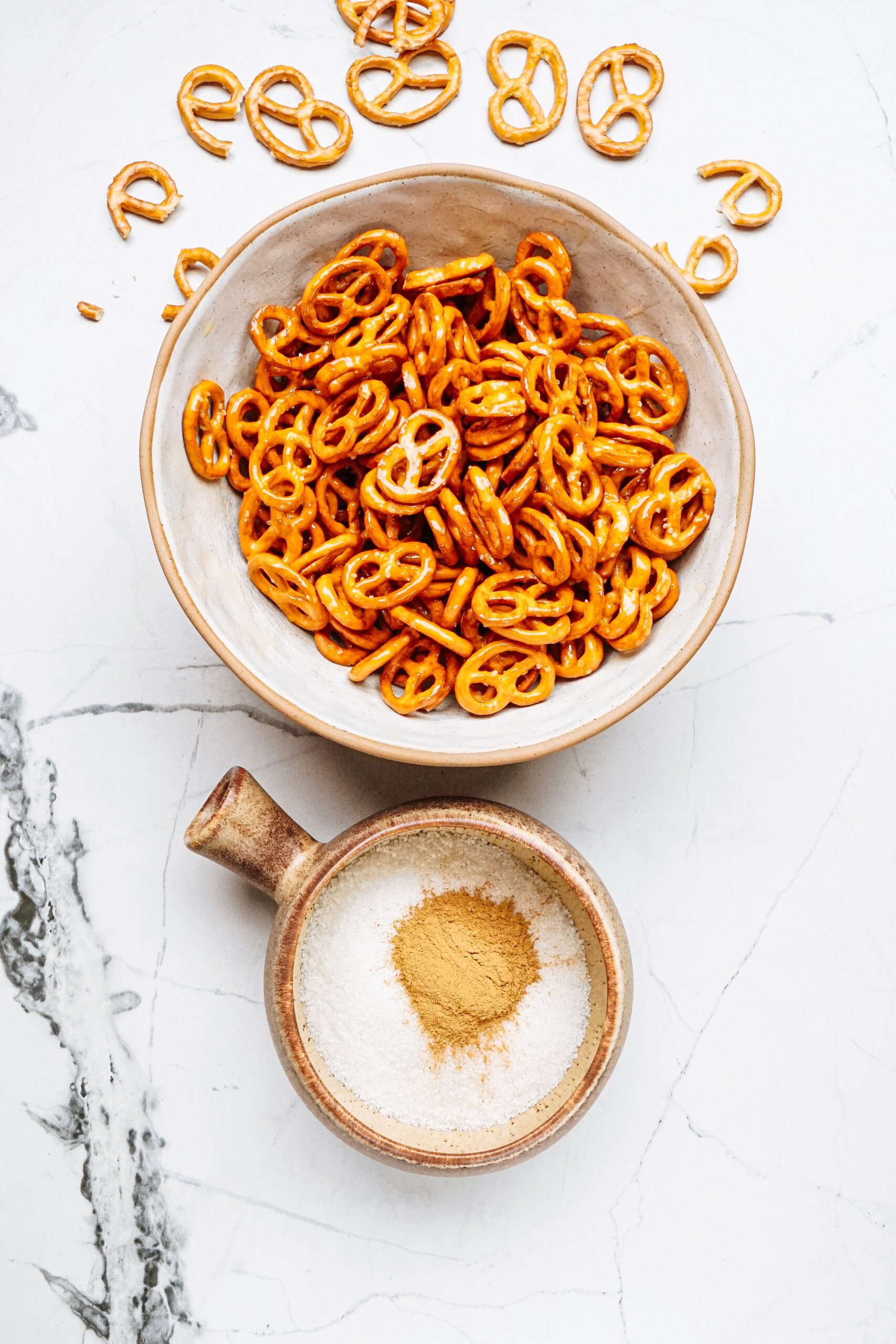 A bowl of cinnamon roll pretzels sits enticingly next to a small dish of white powder dusted with brown spice, all set elegantly on a pristine white marble surface.