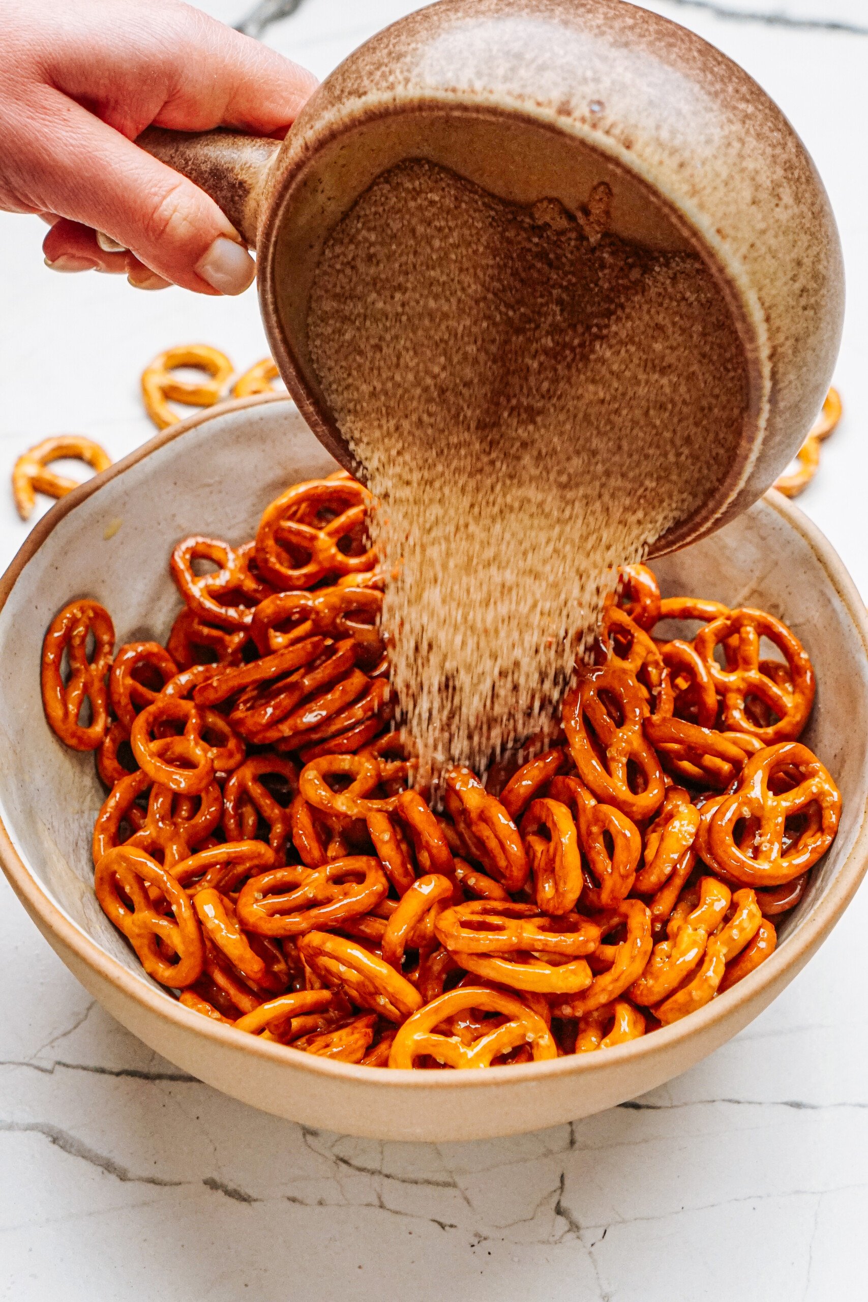 A hand sprinkles sugar over a bowl of cinnamon roll pretzels on a marble surface.