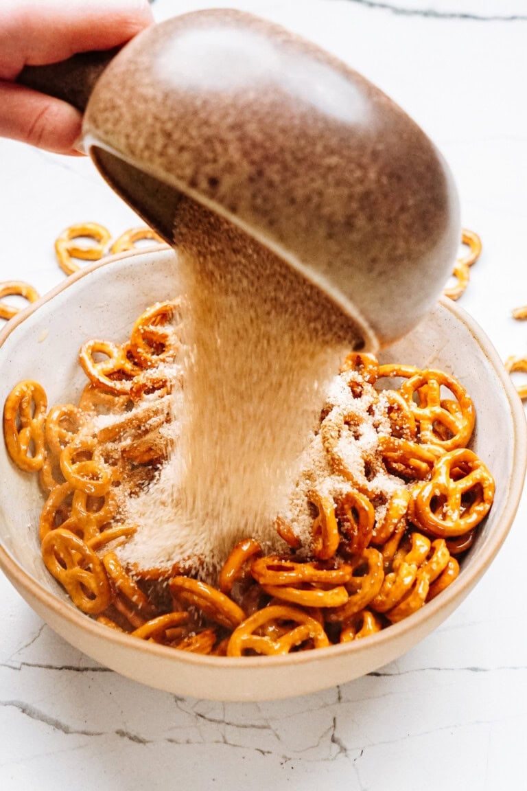 A hand sprinkles powdered seasoning from a brown bowl onto a larger bowl filled with cinnamon roll pretzels on a white marble surface.