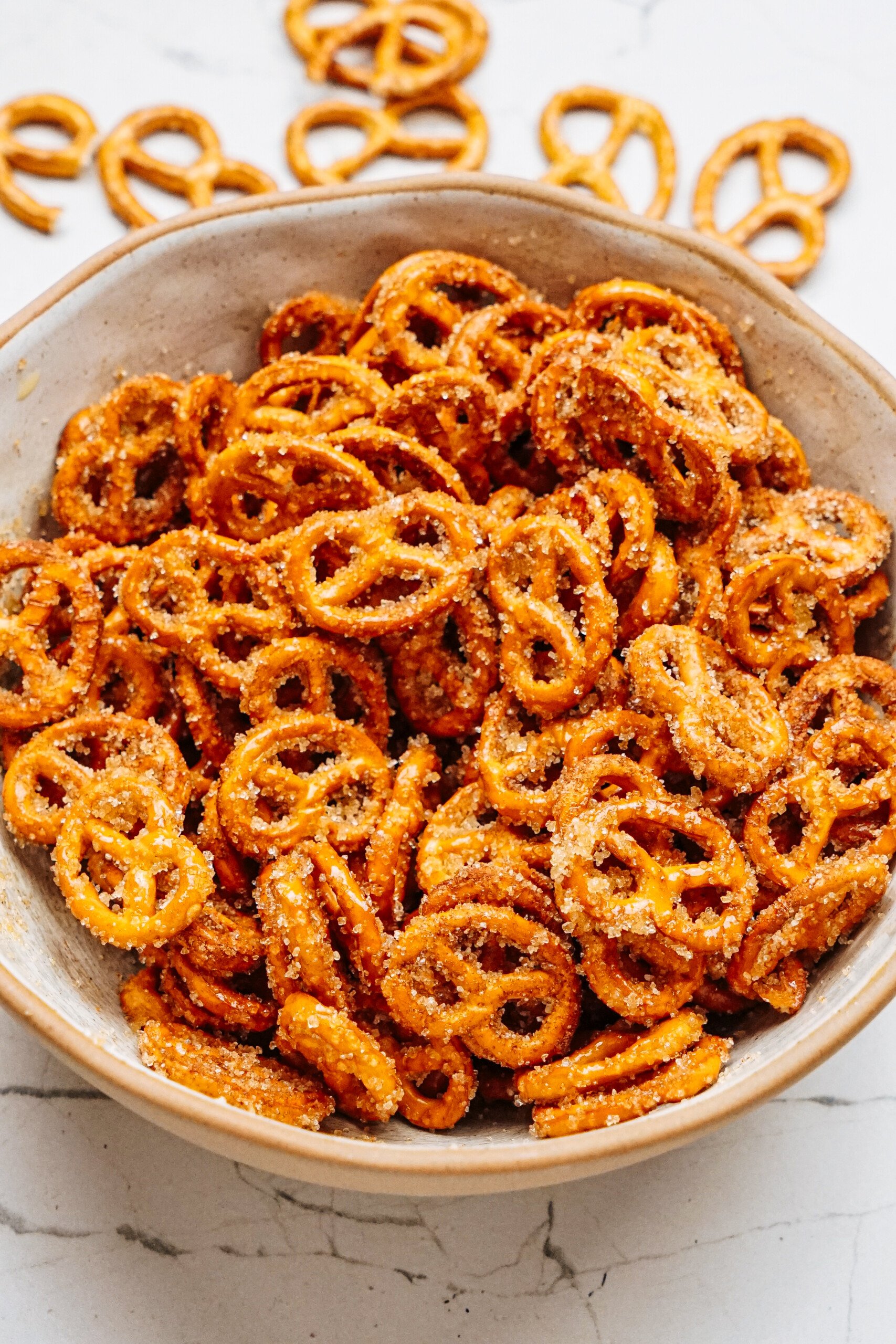 A bowl filled with seasoned mini pretzels, reminiscent of cinnamon roll pretzels, sits on a marble surface, with some scattered nearby.