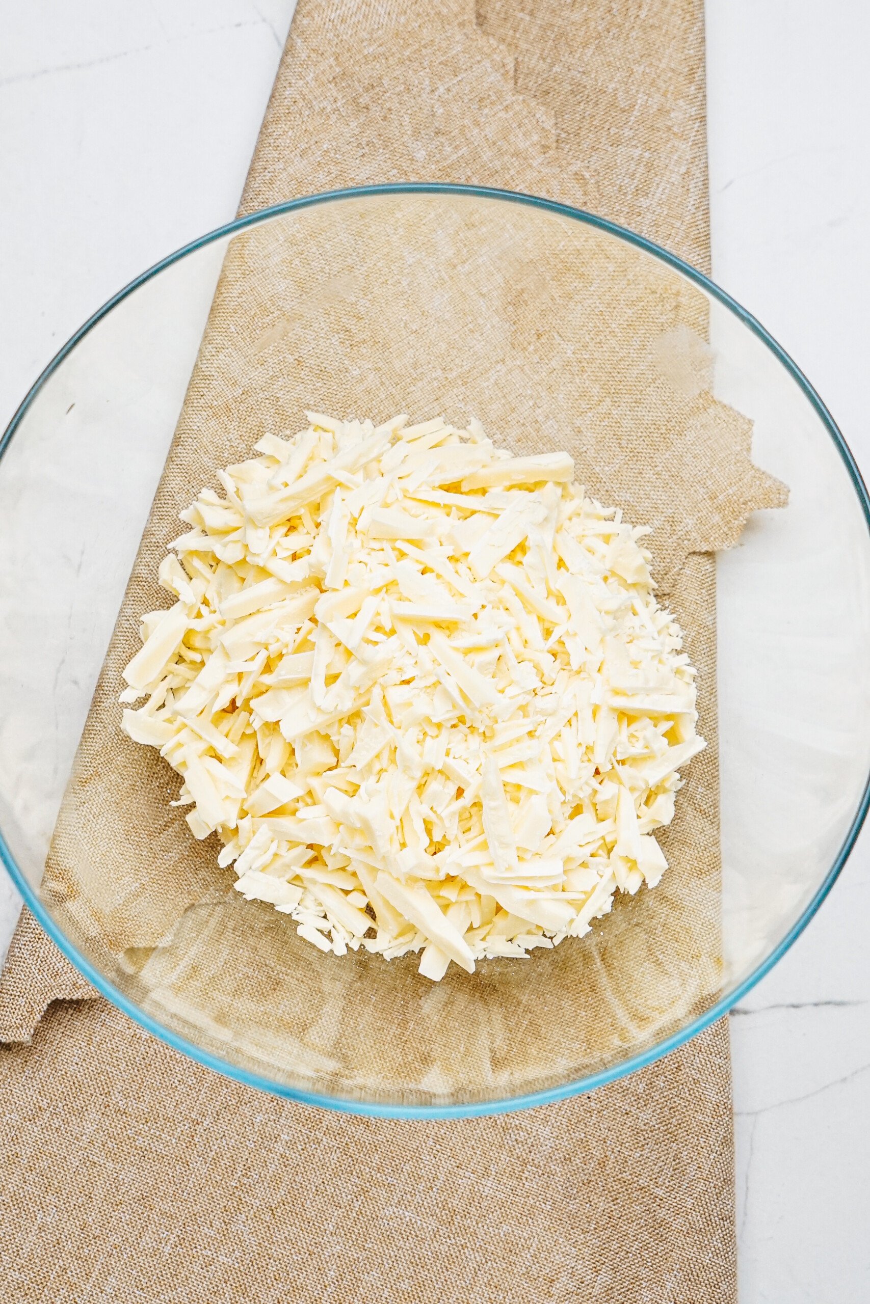 A glass bowl filled with shredded white chocolate is placed on a beige fabric surface, reminiscent of the warm tones of cinnamon roll pretzels.