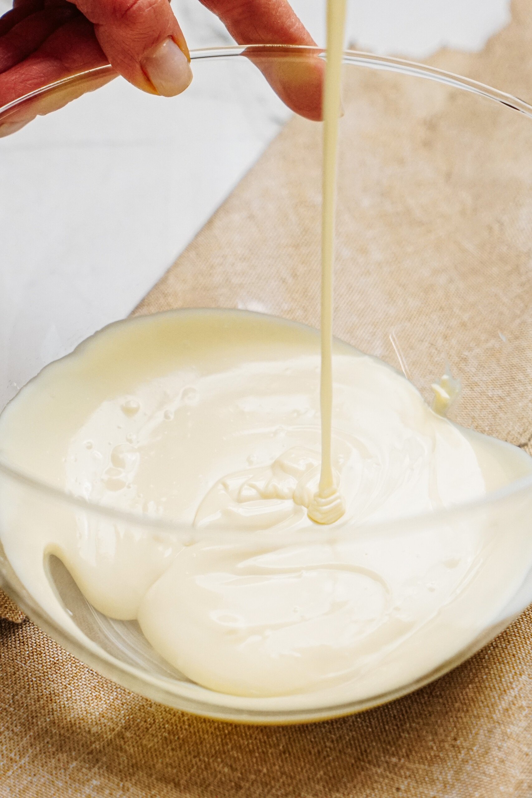 A hand pours a thick, creamy liquid into a clear glass bowl on a burlap surface, evoking the comforting aroma of cinnamon roll pretzels.