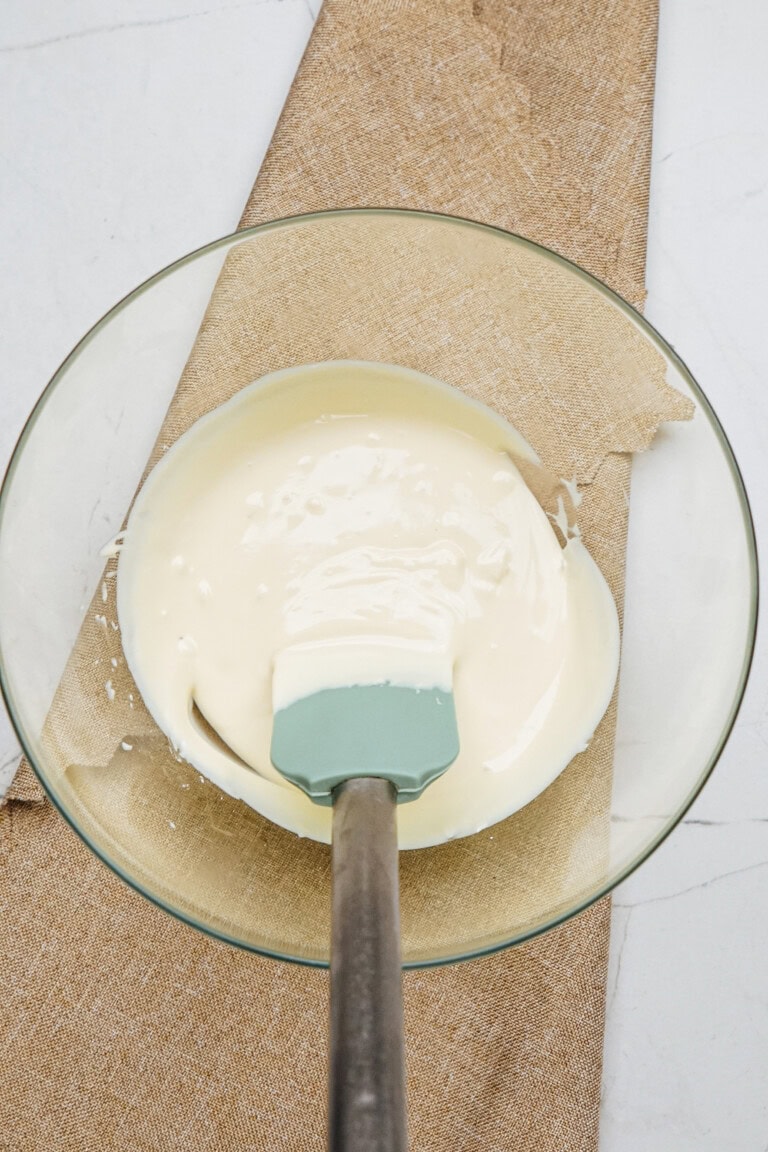 A glass bowl filled with white batter for cinnamon roll pretzels and a spatula rests on a beige cloth.