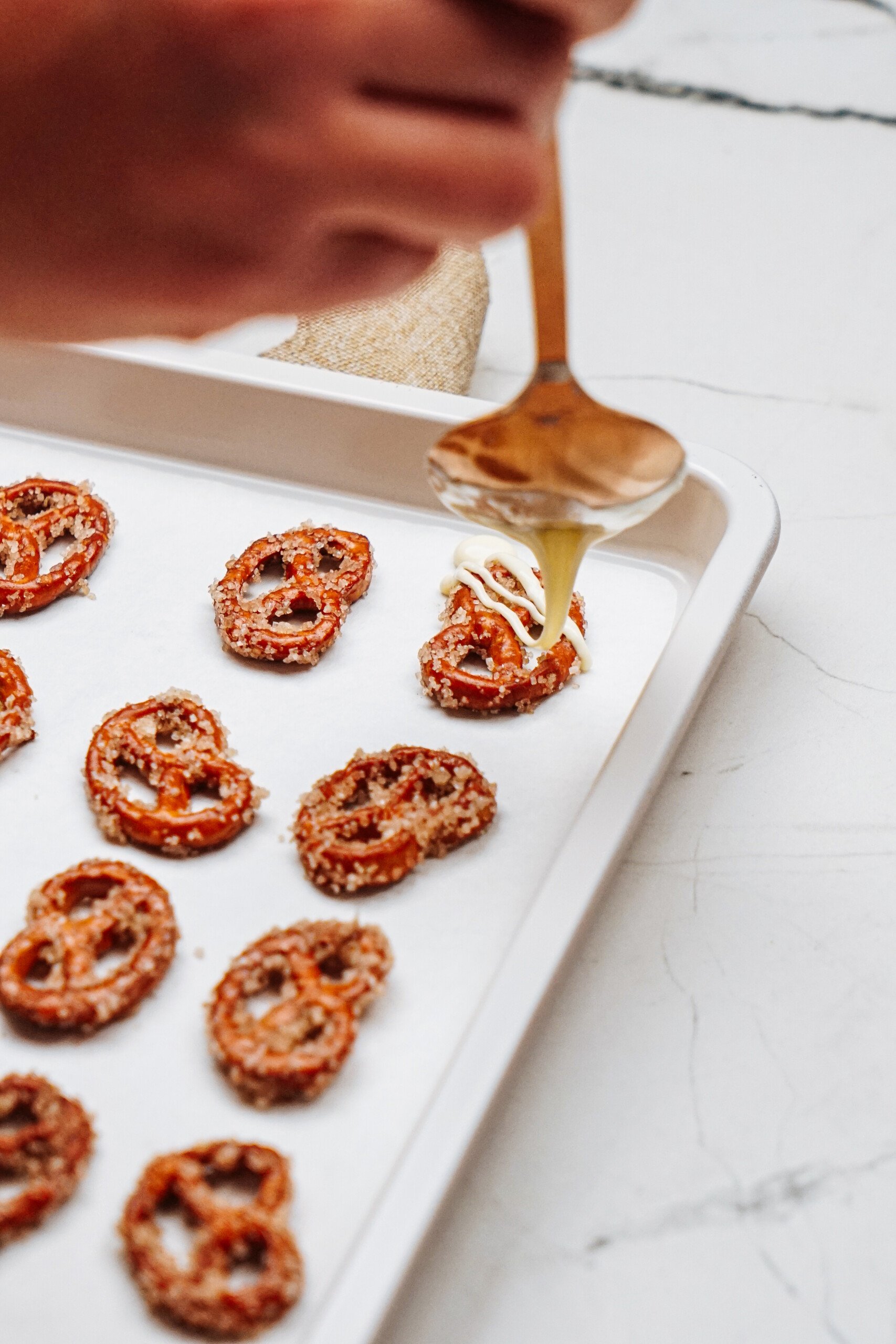 A hand drizzles a light sauce over cinnamon roll pretzels on a baking tray lined with parchment paper.