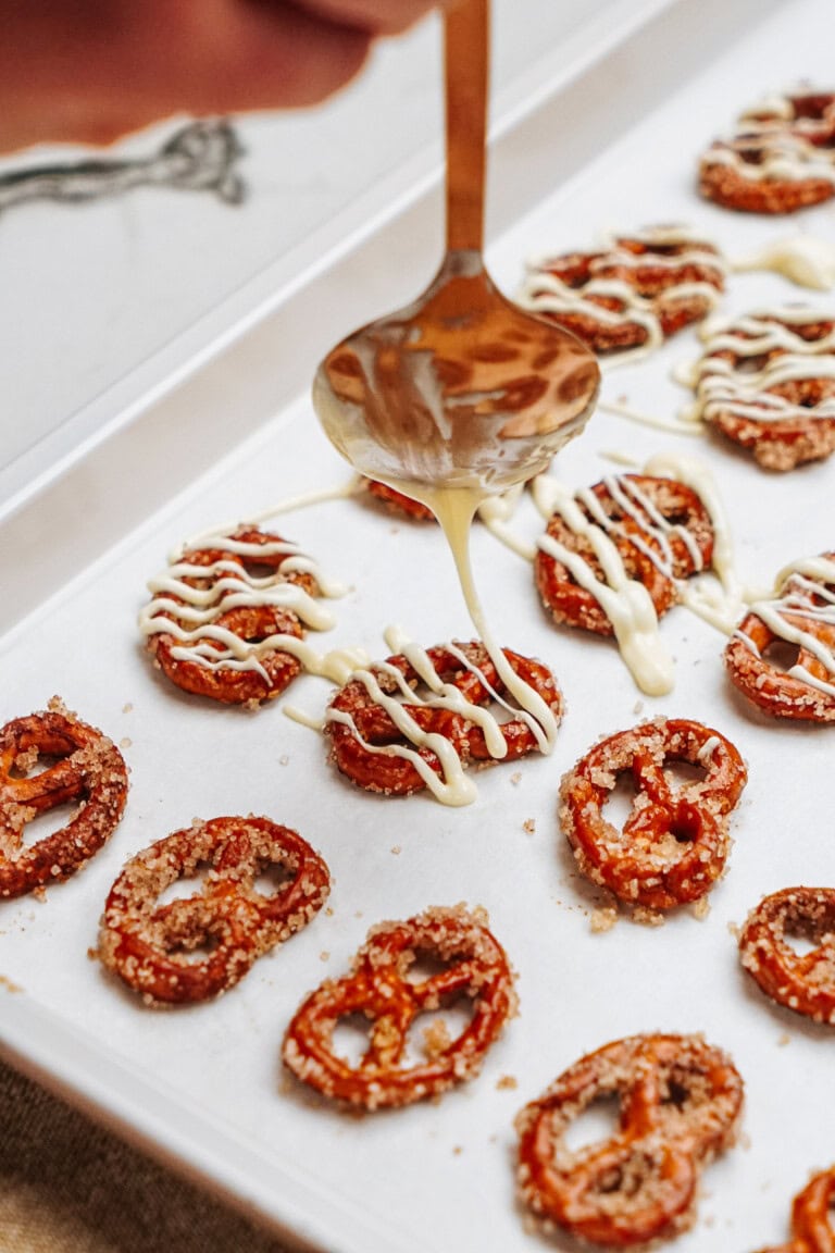 A spoon drizzles white chocolate over cinnamon roll pretzels on a tray.