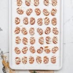 A baking tray of round cinnamon roll pretzels drizzled with white and dark chocolate in neat rows rests on a white marble surface.