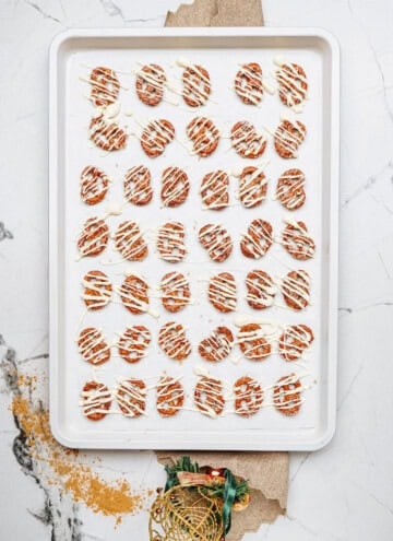 A baking tray of round cinnamon roll pretzels drizzled with white and dark chocolate in neat rows rests on a white marble surface.