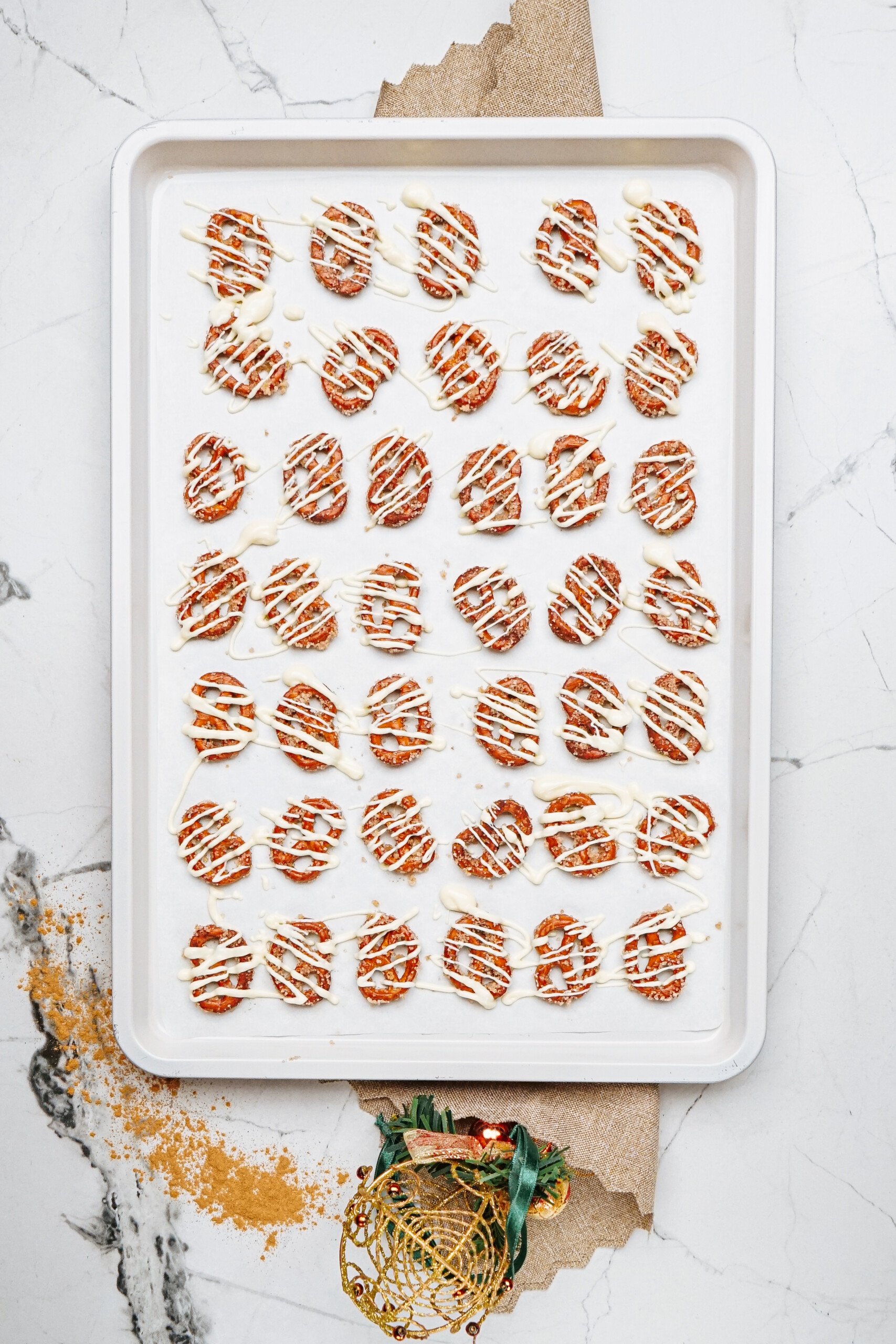 A baking tray of round cinnamon roll pretzels drizzled with white and dark chocolate in neat rows rests on a white marble surface.