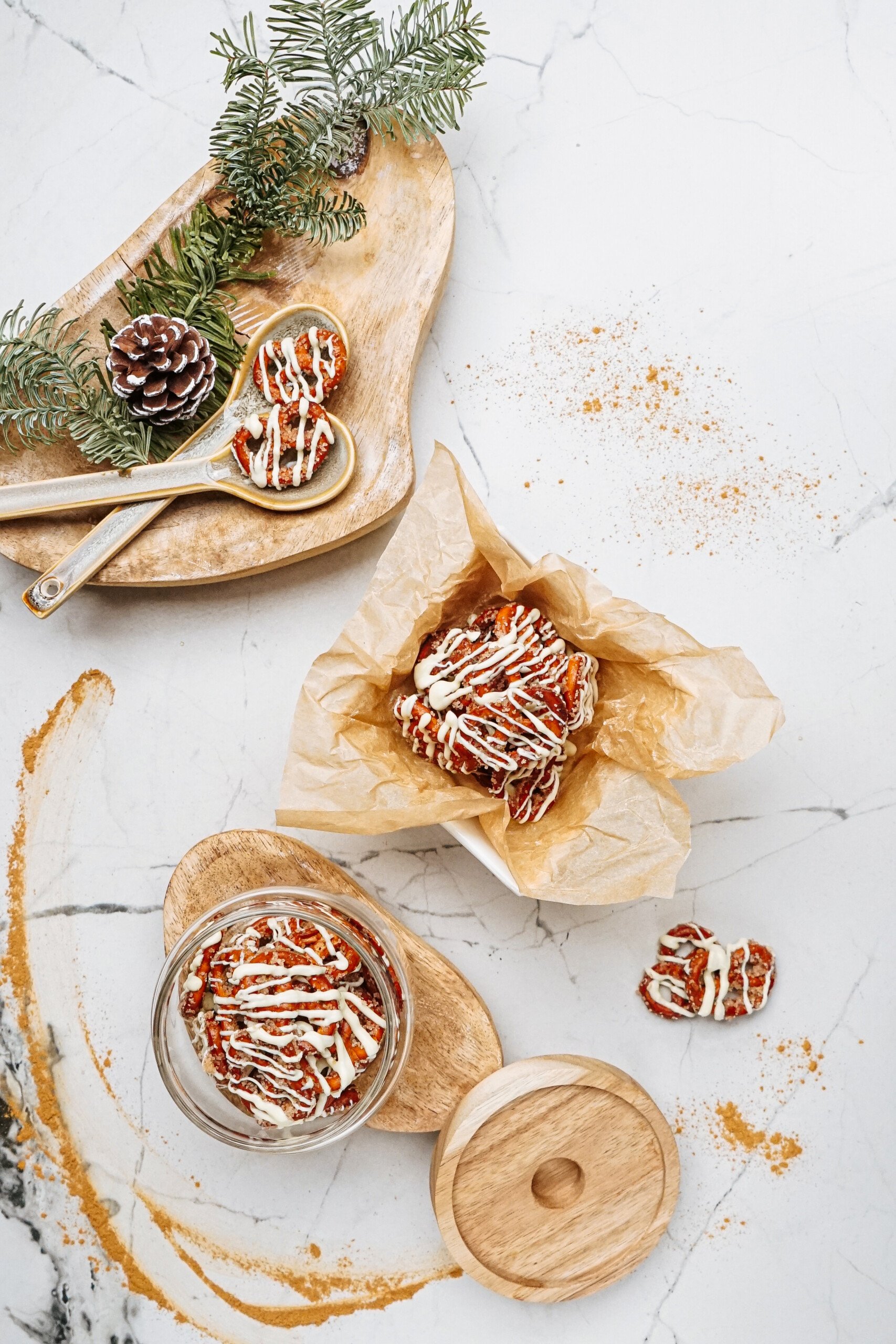 A display of frosted cookies and cinnamon roll pretzels on a wooden board with pinecones and greenery, placed on a marble surface.