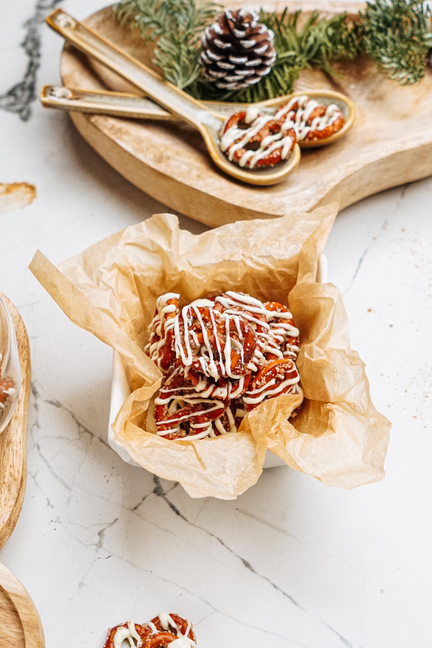 A bowl of cinnamon roll pretzels drizzled with white chocolate sits on parchment paper, surrounded by festive decor of pinecones and greenery.