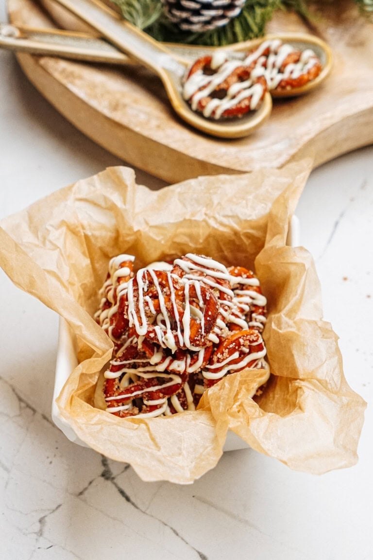 A bowl lined with wax paper holds cinnamon roll pretzels drizzled with white icing, reminiscent of cinnamon roll pretzels, placed on a marble counter with a wooden board and decorative spoons in the background.