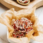 A bowl of cinnamon roll pretzels drizzled with white icing sits on crinkled parchment paper, with wooden spoons of nuts resting in the background on a rustic wooden tray.