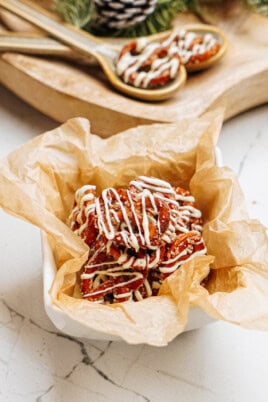 A bowl of cinnamon roll pretzels drizzled with white icing sits on crinkled parchment paper, with wooden spoons of nuts resting in the background on a rustic wooden tray.