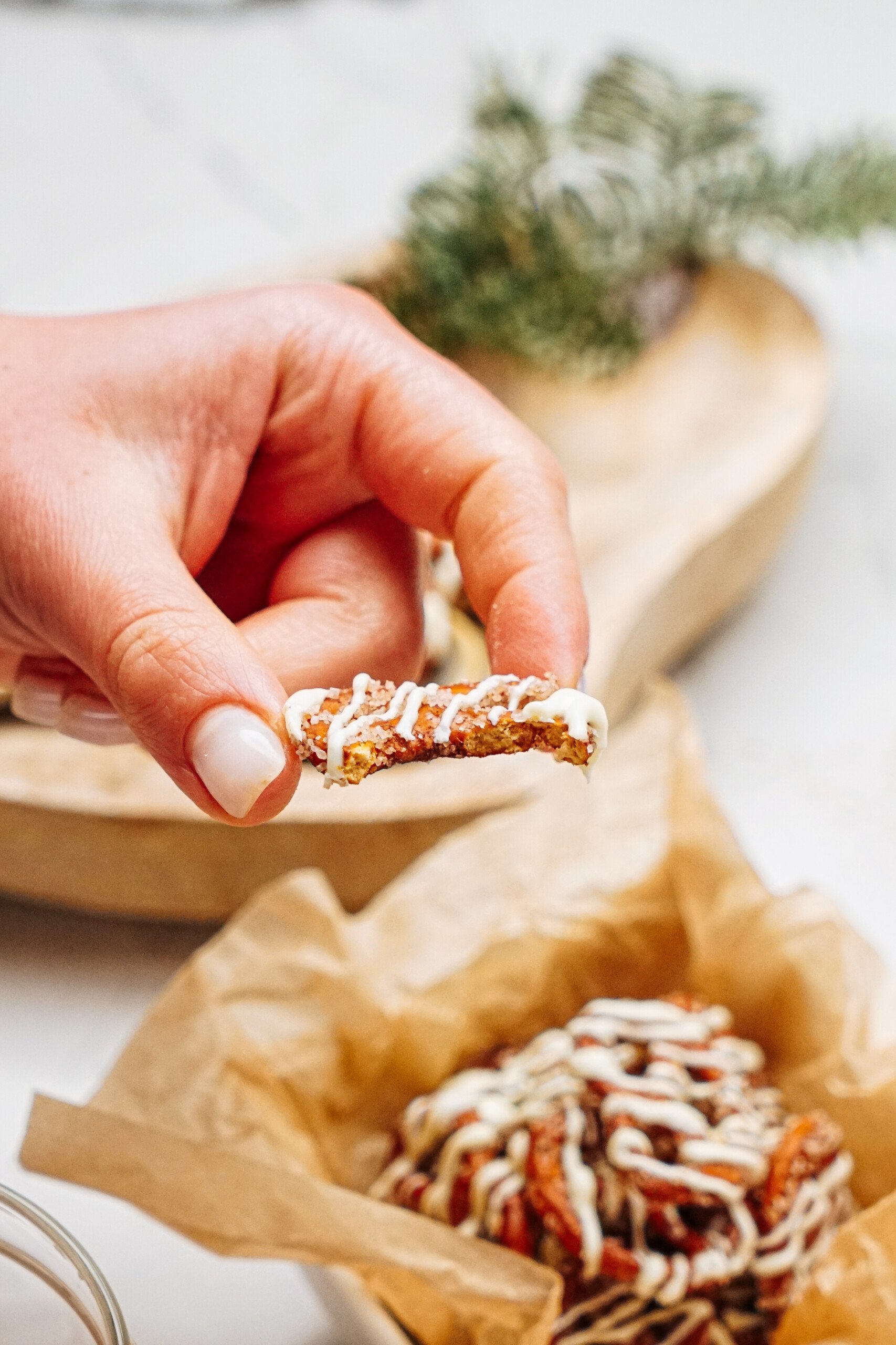 A hand holds a small piece of cinnamon roll drizzled with white icing. A wooden tray with more pastries and pretzels is in the background.