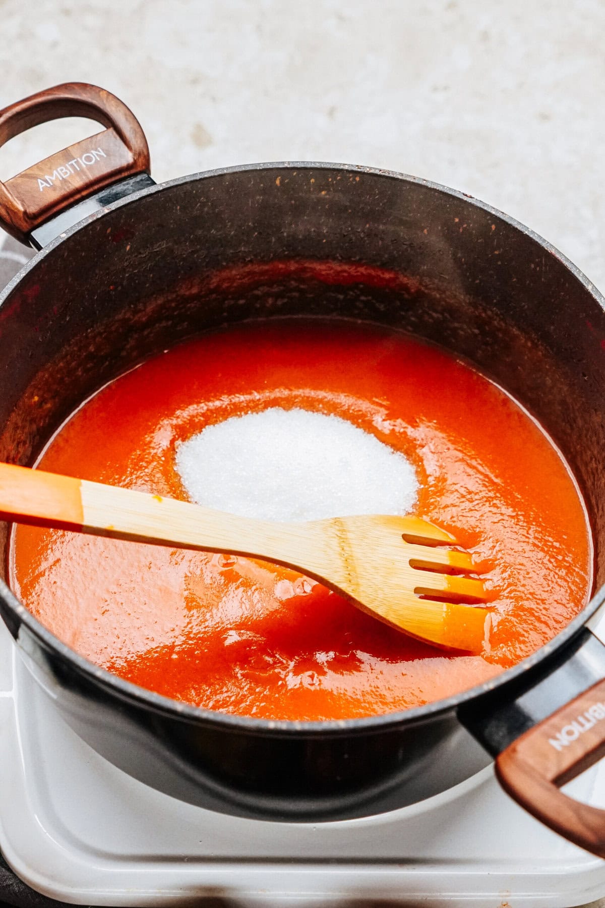 A black pot filled with red tomato sauce simmers on the stove, preparing for a delicious batch of stuffed peppers. A wooden spoon rests inside, gently coaxing the flavors together, while a mound of sugar sits sweetly atop the simmering mixture.