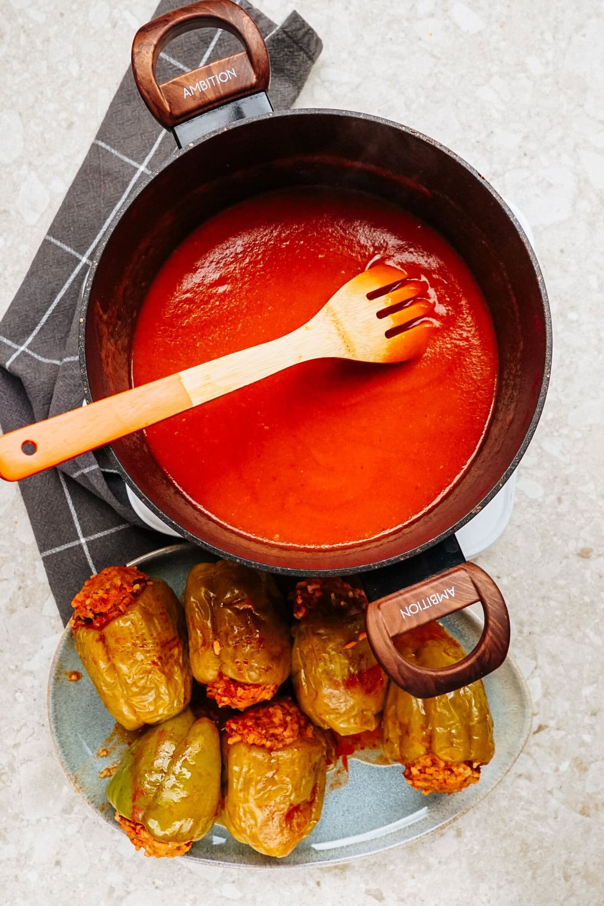 A pot of tomato sauce with a wooden spoon, next to a plate of savory stuffed peppers on a checkered cloth.