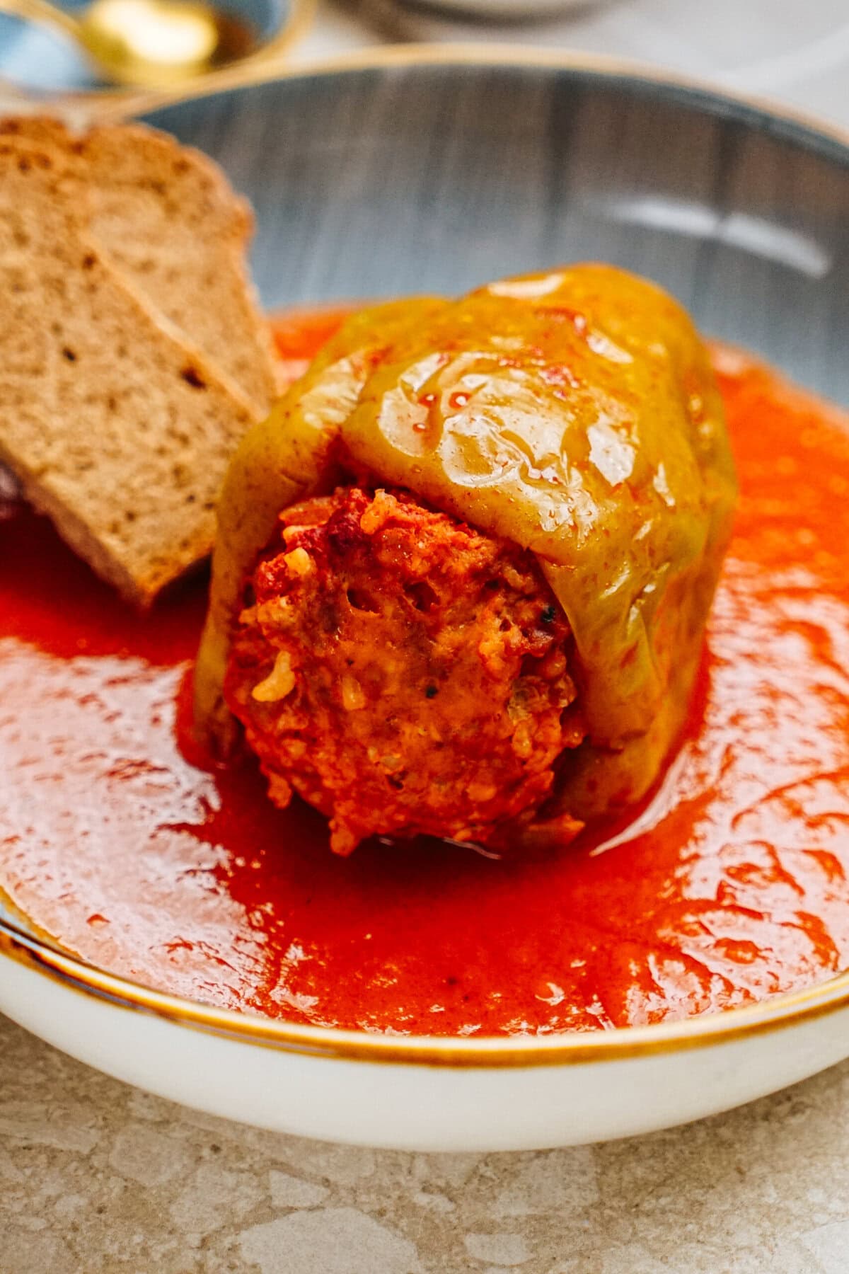 Savor the delicious stuffed peppers, brimming with rice and meat in rich tomato sauce, served in a bowl alongside a slice of hearty brown bread.