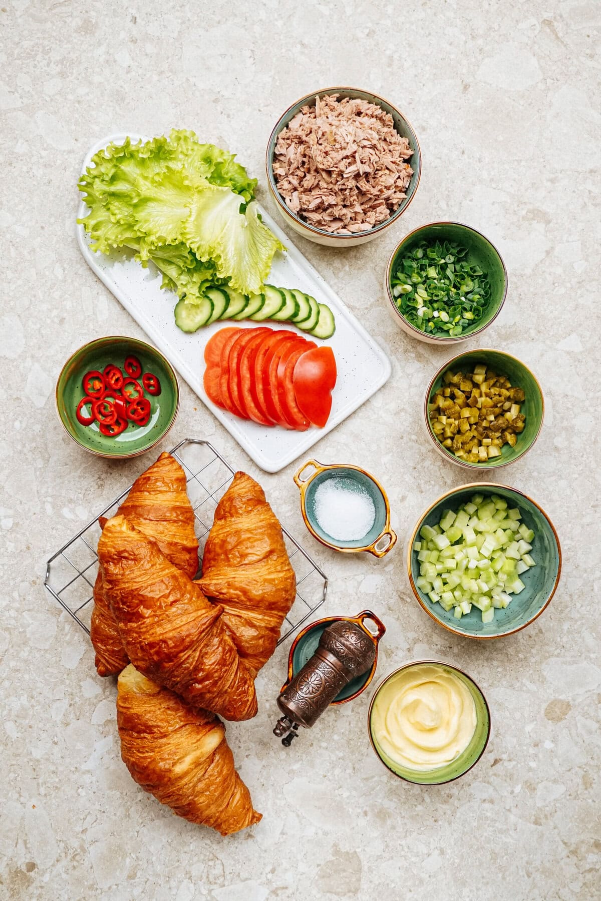 A delectable array of ingredients for a tuna salad sandwich, featuring flaky croissants, fresh sliced vegetables, creamy mayonnaise, and savory tuna, artfully arranged on a marble surface.