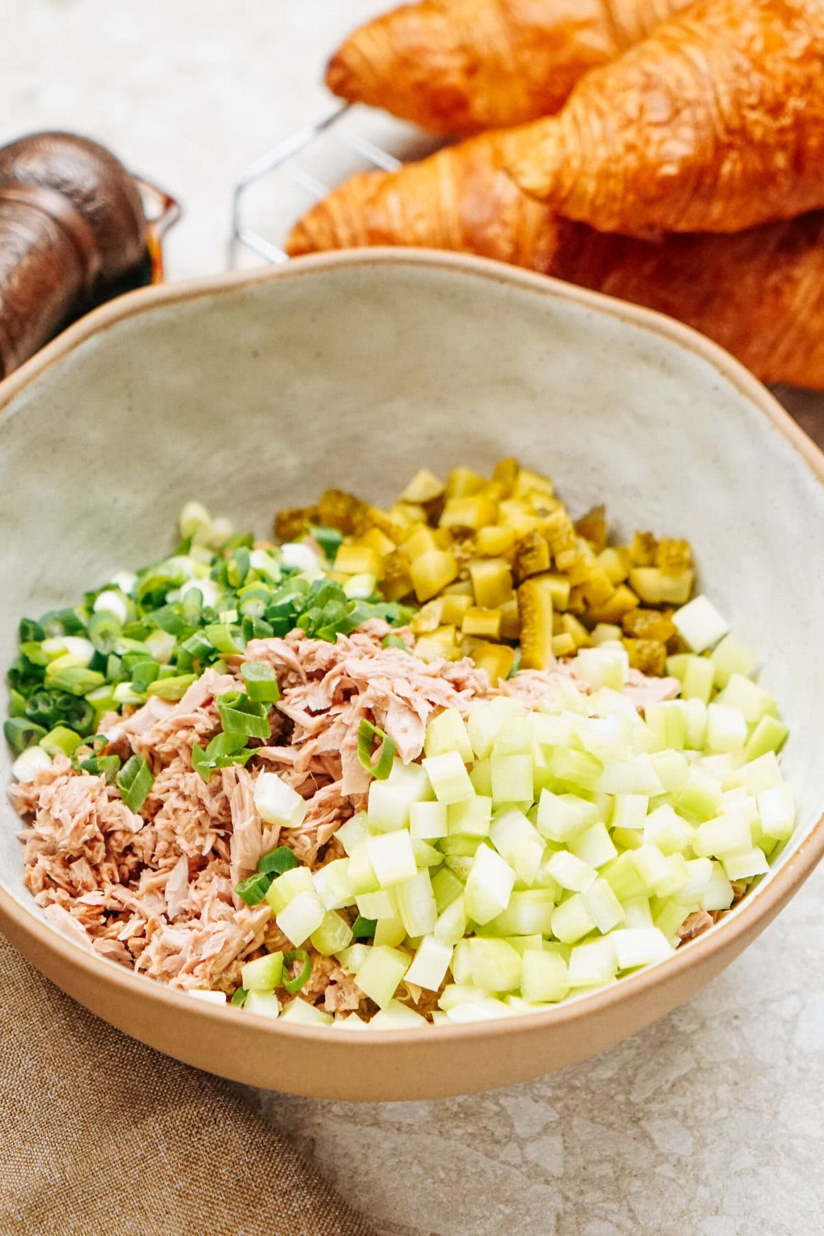 A bowl featuring a mix of chopped tuna, pickles, green onions, and cucumber captures the essence of a deconstructed Tuna Salad Sandwich, perfectly complemented by croissants in the background.