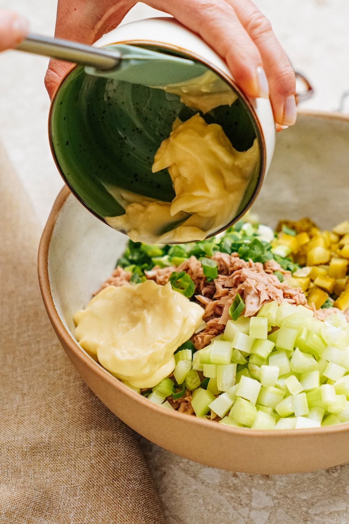 Mayonnaise is being poured from a green bowl into a larger bowl containing tuna, chopped celery, and pickles, creating the perfect mix for a delicious tuna salad sandwich.