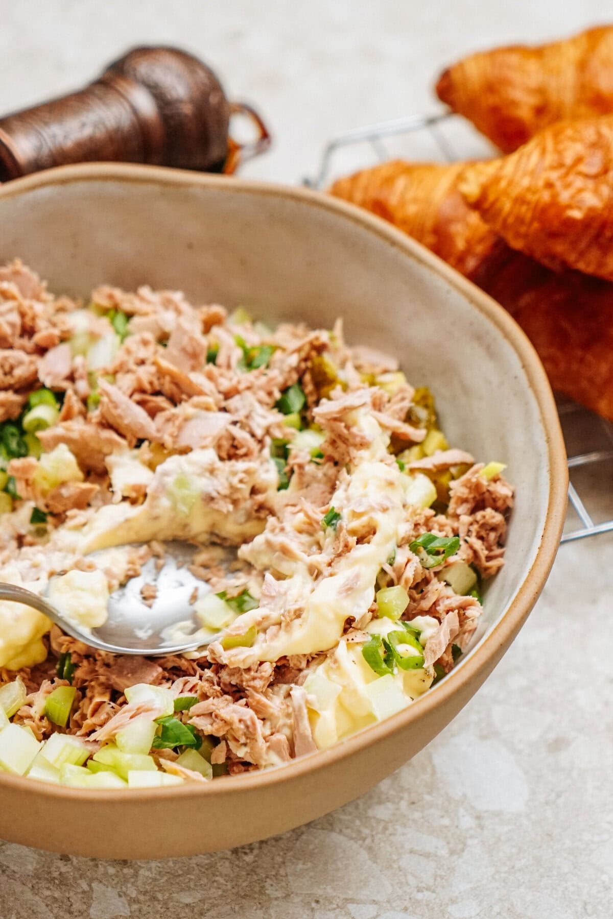 A bowl of tuna salad with green onions and mayonnaise sits beside fresh croissants, perfect for crafting a delicious tuna salad sandwich, all on a sleek marble surface with a pepper grinder nearby.