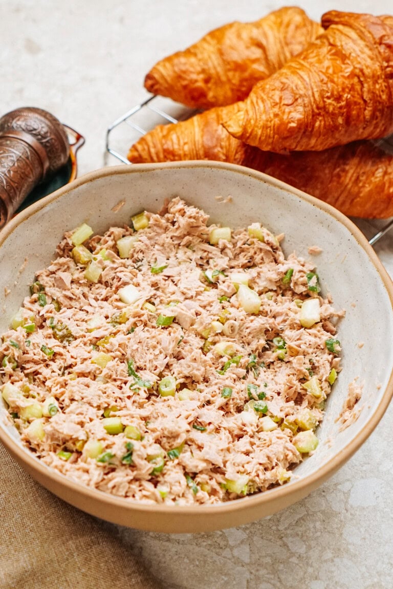 A bowl of tuna salad with diced celery sits on a table, ready to fill freshly baked croissants for a delightful tuna salad sandwich. Nearby, a pepper grinder awaits to add the perfect final touch.