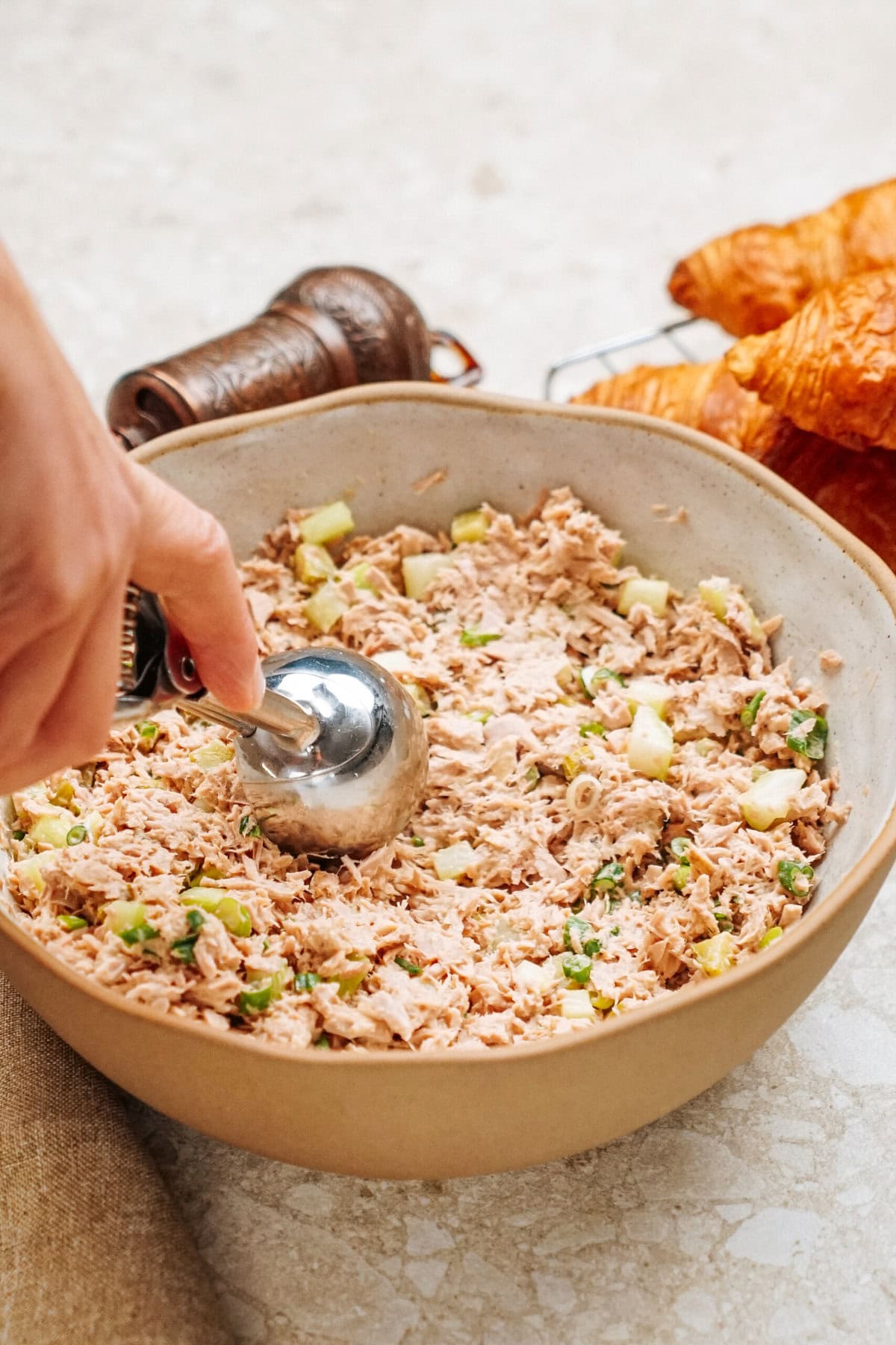 A hand using a scooper serves tuna salad with chopped ingredients from a bowl, ready to create the perfect tuna salad sandwich. Croissants and a pepper grinder linger invitingly in the background.